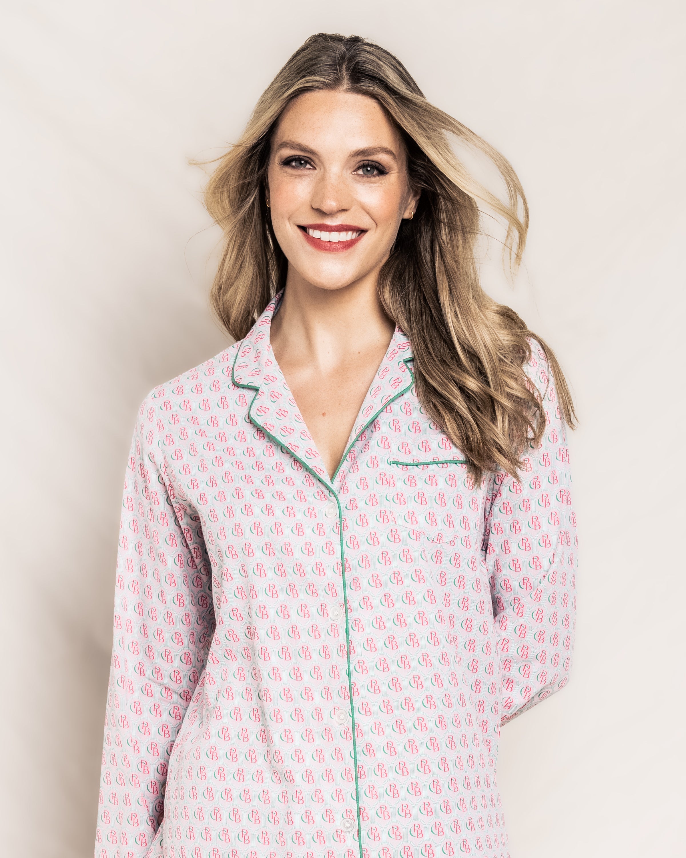 A smiling woman with long blonde hair wears a Colony Hotel x Petite Plume Womens Exclusive Print Pajama Set by Petite Plume. She stands against a light background, slightly tilting her head, with her hair gently flowing to the side.