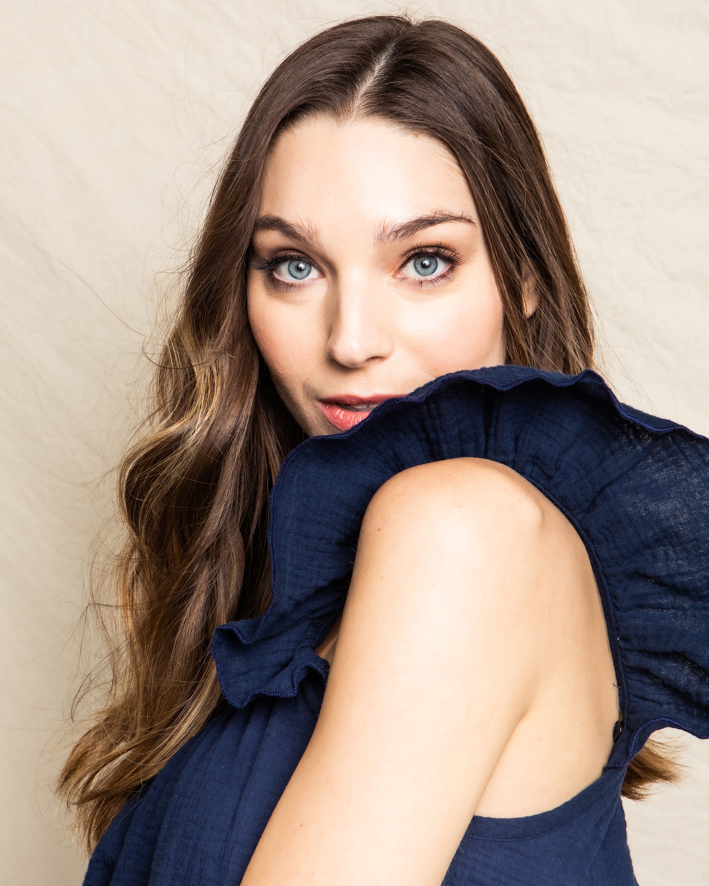 A woman with long, wavy brown hair and striking blue eyes wears the Petite Plume Womens Gauze Celeste Top in Navy. The luxury cotton gauze and ruffled sleeves enhance her feminine silhouette as she looks over her shoulder at the camera against a light background.