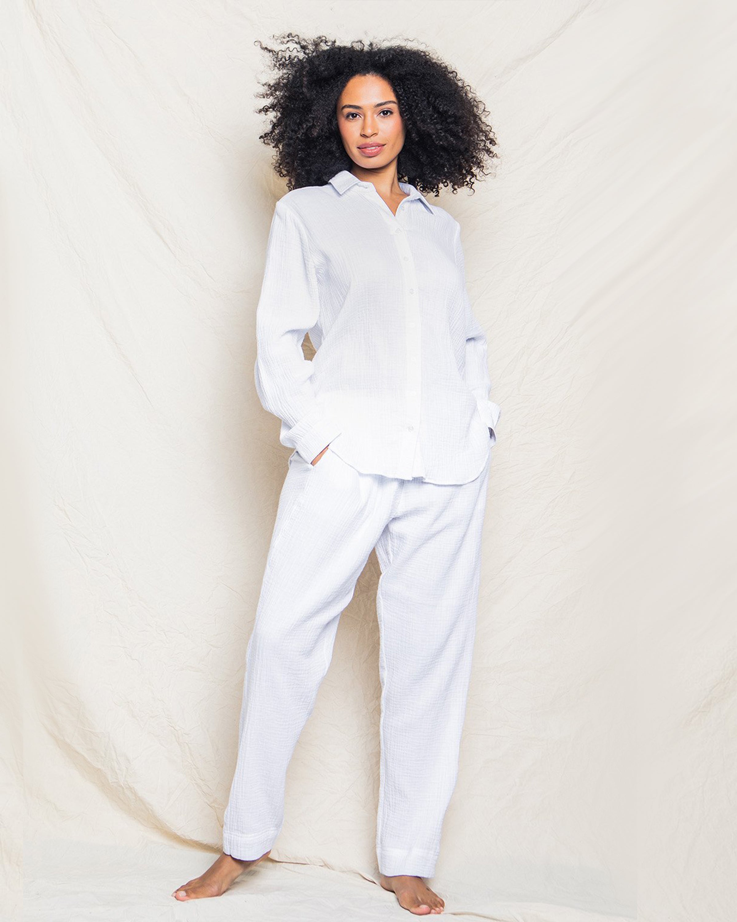 A person with curly hair stands barefoot on a neutral background wearing Petite Plumes Womens Gauze Drawstring Pants in white, crafted from soft cotton gauze. With hands in pockets, the long-sleeve button-up shirt and gauze pants create a relaxed, comfortable look.