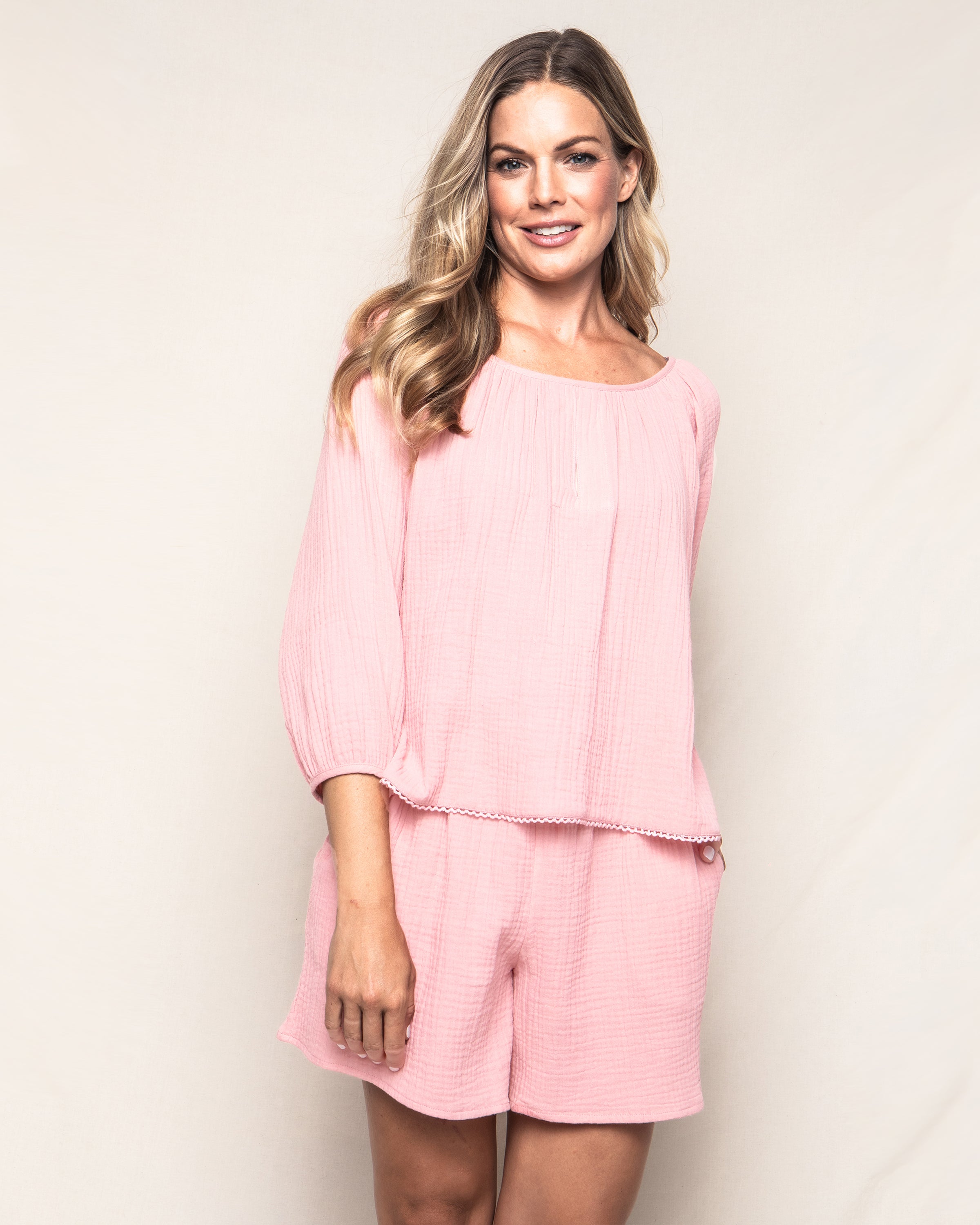 A person smiling in Petite Plume Womens Gauze Drawstring Shorts in pink and a matching light pink top. The outfit, crafted from soft cotton gauze, complements their long, wavy blonde hair as they pose confidently against a plain background.