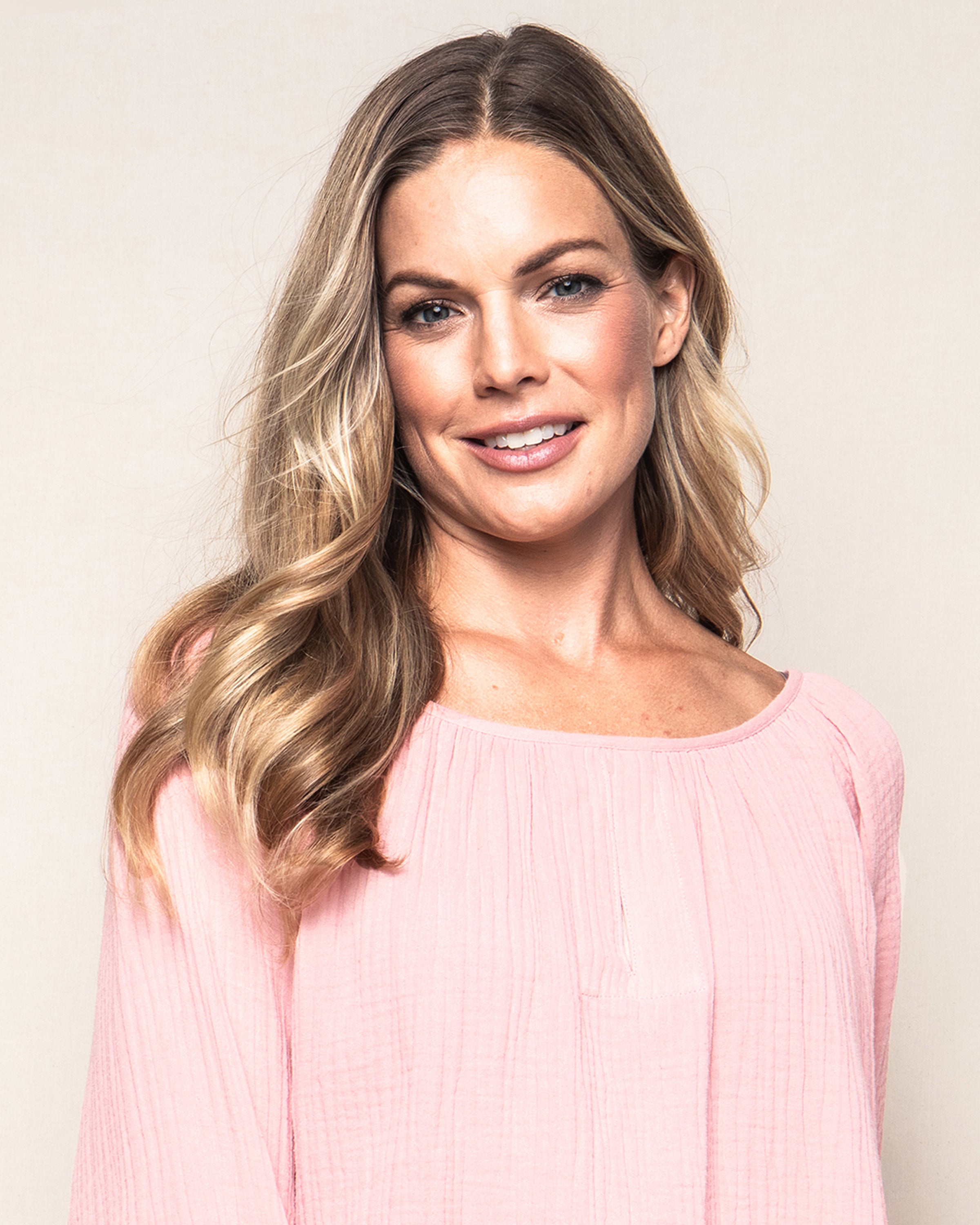 A woman with long, wavy blonde hair wears a light pink Womens Gauze Willow Scoopneck Top by Petite Plume and smiles at the camera against a plain background.