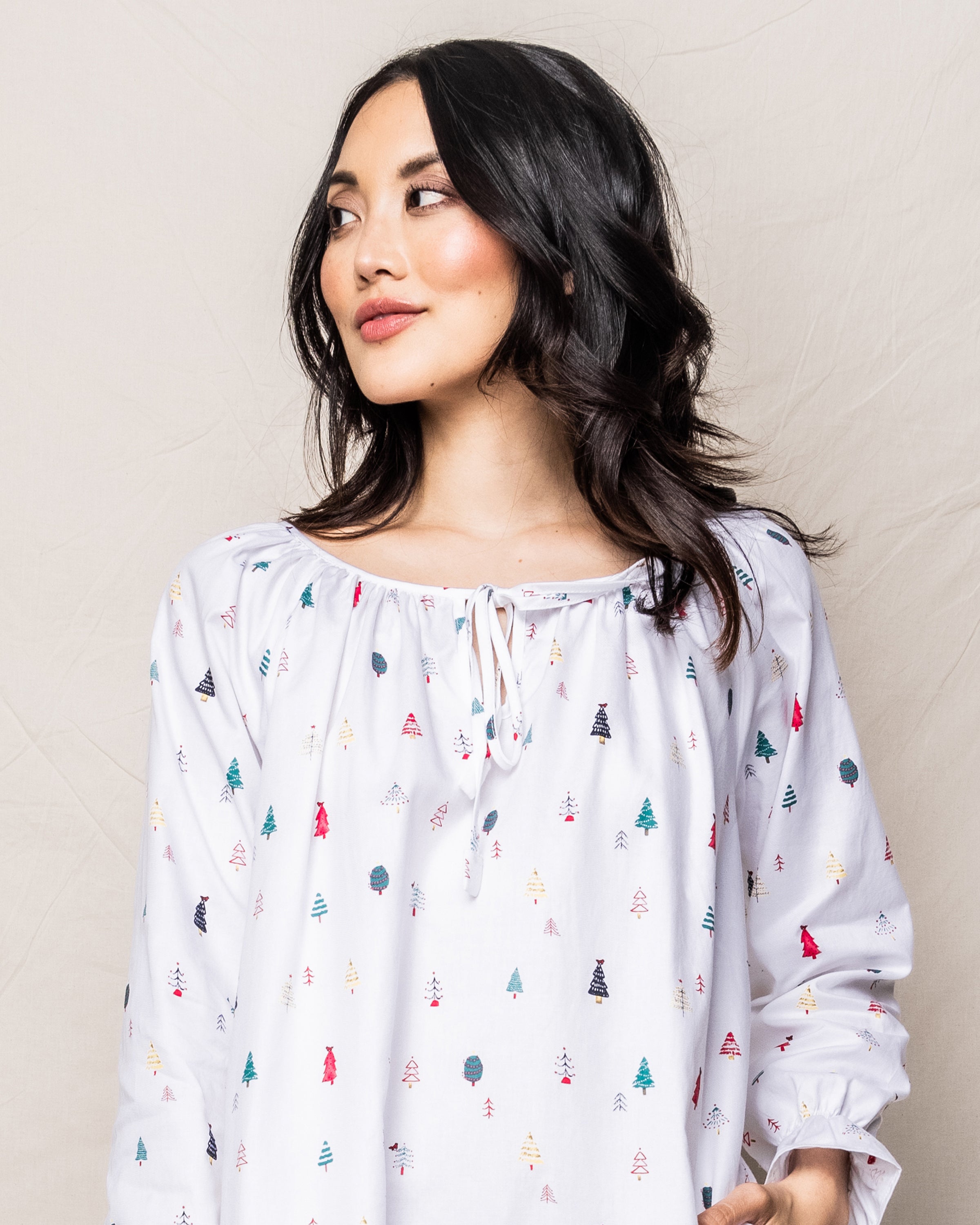 A woman with long dark hair wears the Womens Twill Delphine Nightgown in Merry Trees by Petite Plume, adorned with colorful Christmas tree prints. She gazes slightly to the side against a light, textured background reminiscent of cozy cotton sleepwear.