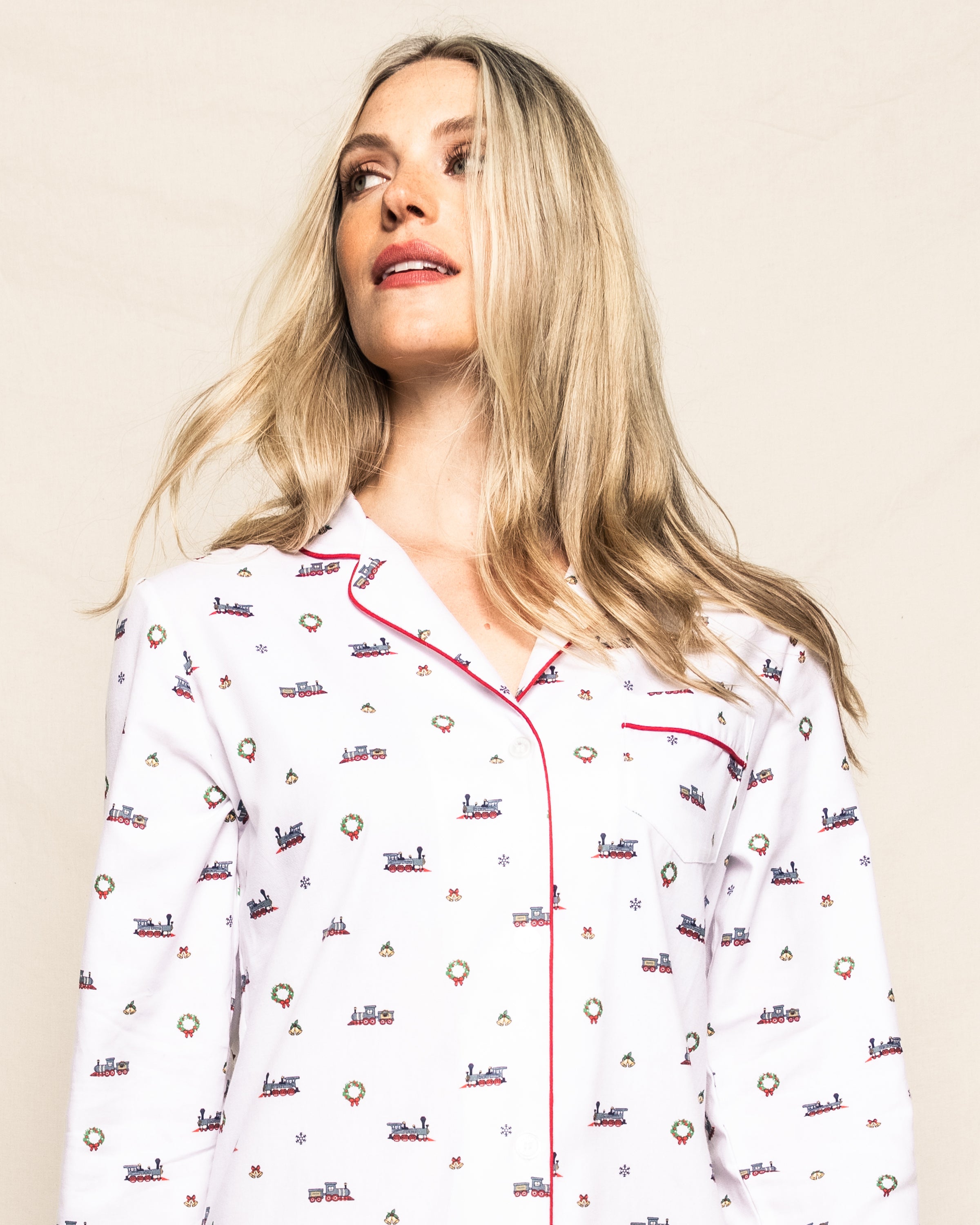 A long-haired person wearing Petite Plumes Womens Twill Nightshirt in Arctic Express style, with train and Christmas prints, and red piping on the collar and pocket. The background is a plain light color.