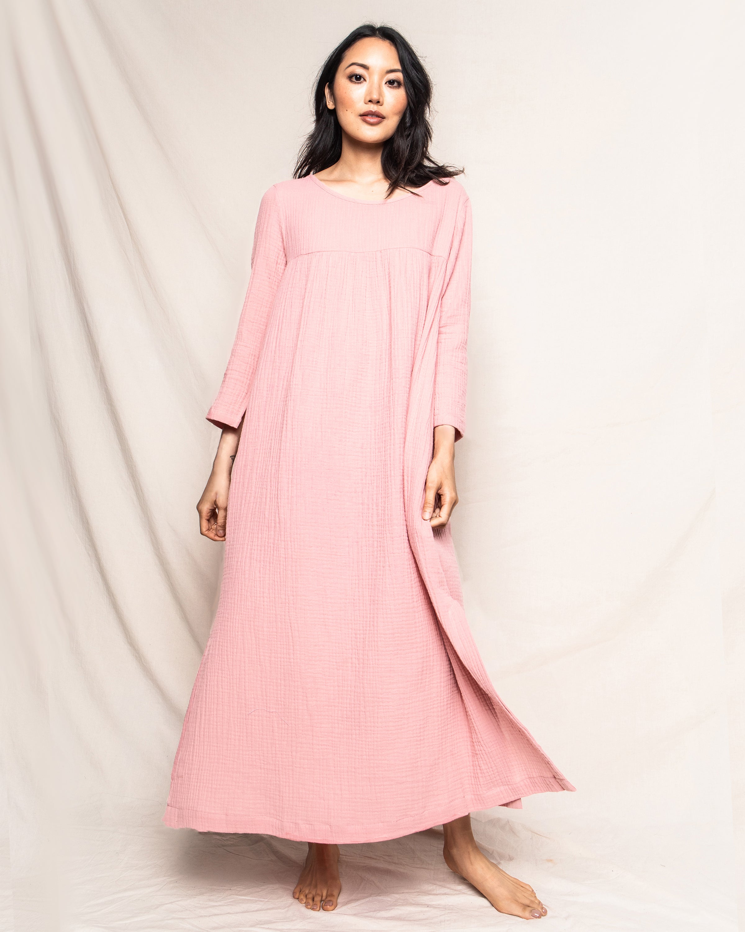 Barefoot and relaxed, a person stands against a plain background wearing the Womens Gauze Provence Sundress in Pink by Petite Plume. Made of lightweight cotton gauze, this long, loose-fitting dress features an elegant design typical of luxury loungewear.