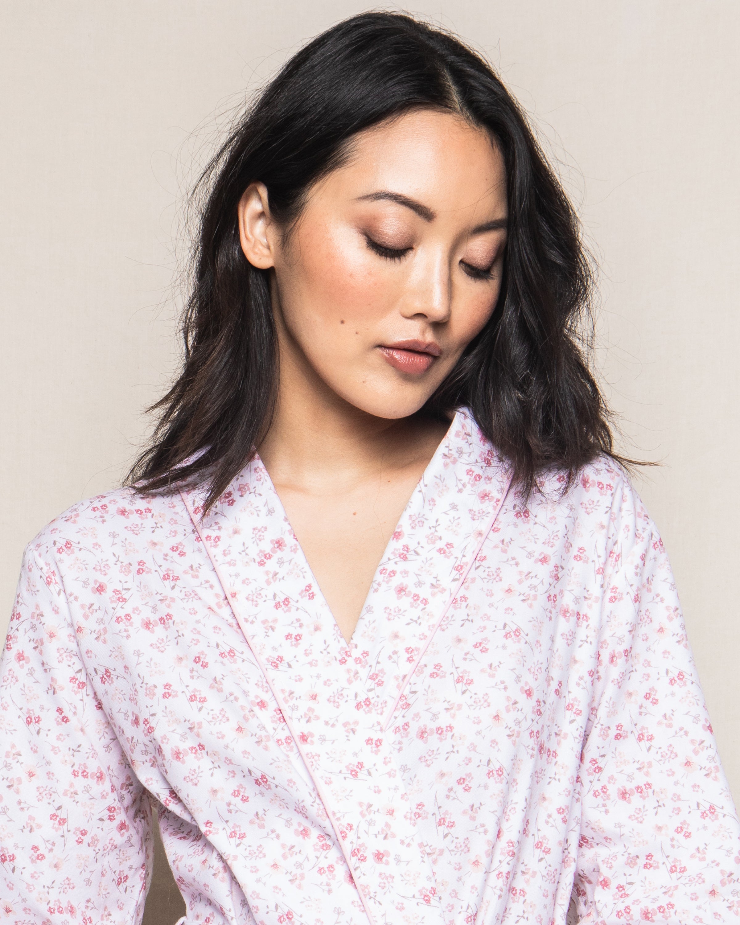 A woman with shoulder-length black hair wears the Womens Twill Robe in Dorset Floral by Petite Plume, featuring an elegant light pink floral design crafted from fine cotton. She gazes down serenely against a neutral backdrop.