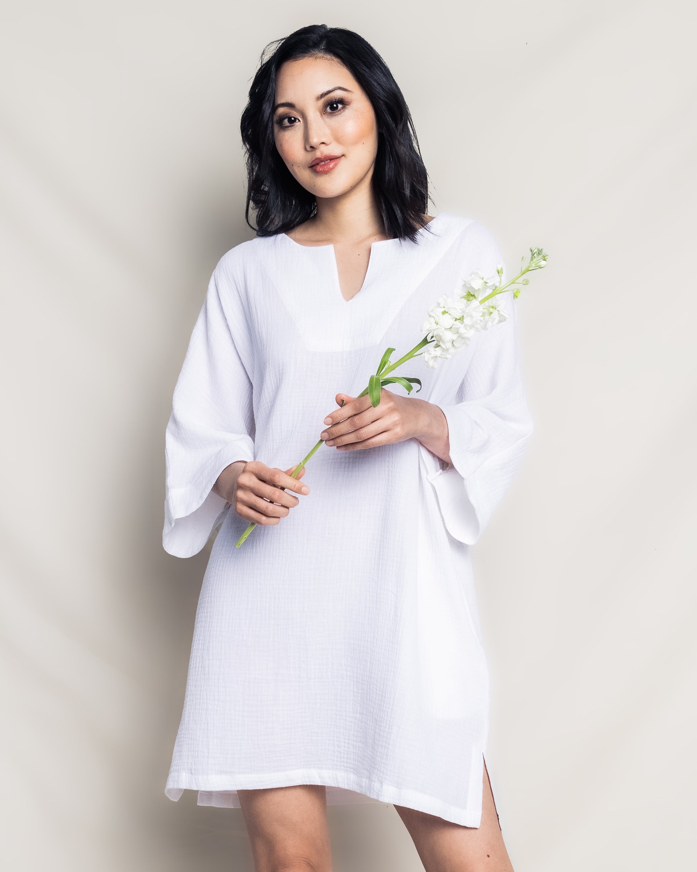 A woman in a Womens Gauze Short Caftan in White by Petite Plume, crafted from luxurious cotton gauze, holds a small white flower bouquet. The dress features loose sleeves and a v-neckline. She has shoulder-length dark hair and stands against a plain backdrop, looking directly at the camera with a slight smile.