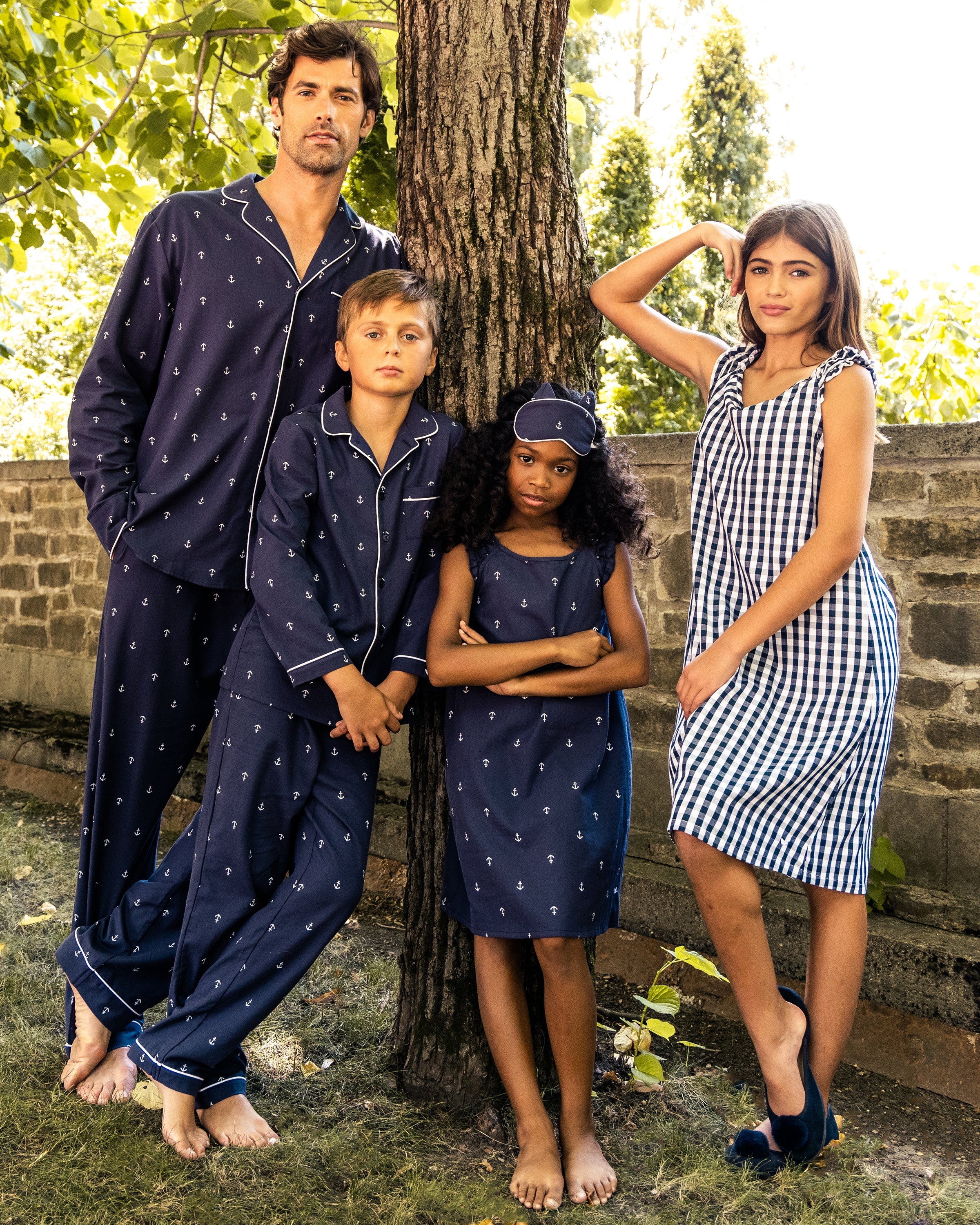 Four people are in a garden with a tree and stone wall backdrop. A man and boy in matching navy pajamas with anchors, a girl in Petite Plumes Amelie Nightgown featuring Portsmouth Anchors print and sleep mask, and another girl in a black-and-white checkered dress stand barefoot on the grass.