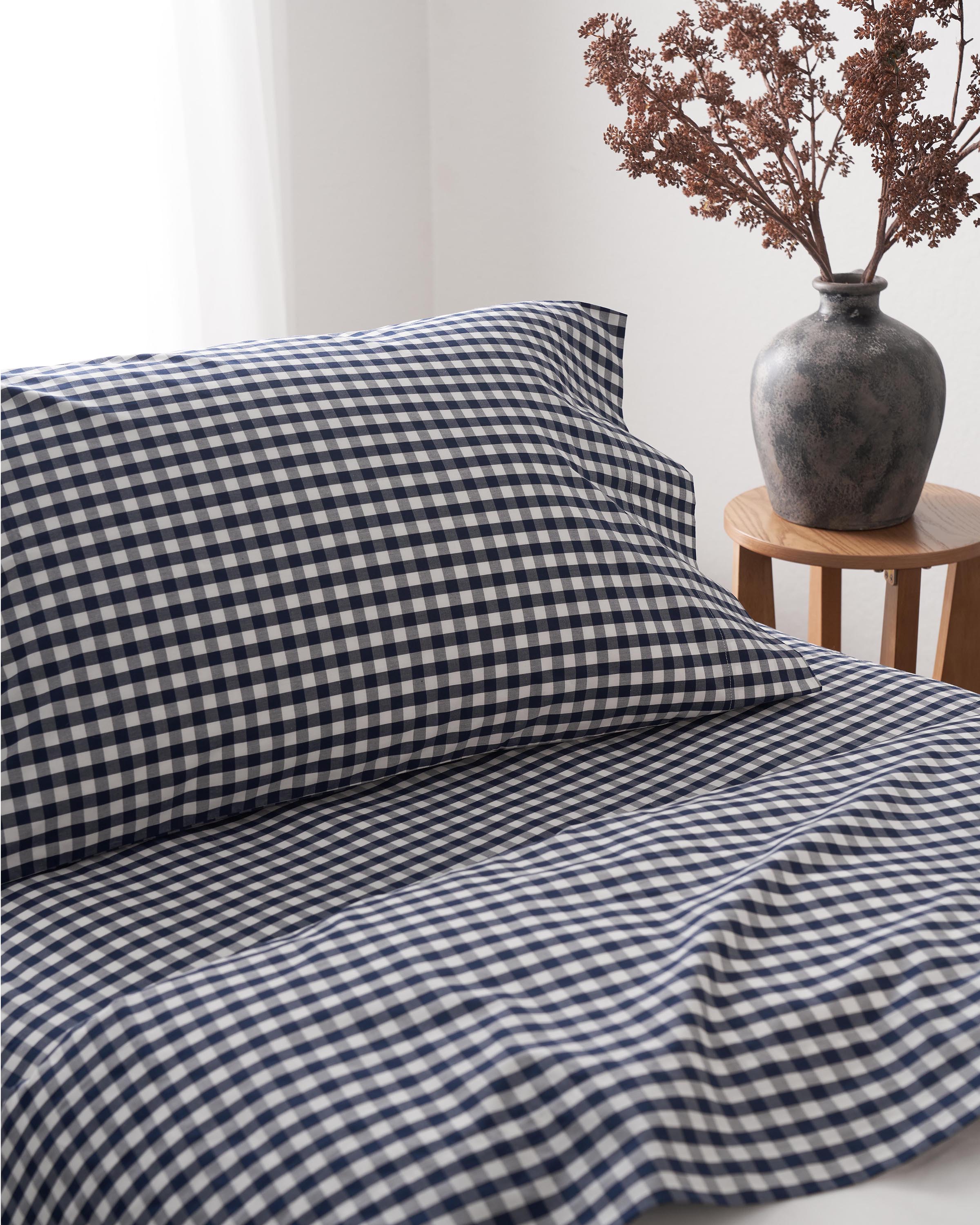 A bed with Luxe Premium Cotton Navy Gingham Bed Sheets by Petite Plume lies invitingly. Nearby, a gray vase with dried foliage sits on a wooden stool. Natural light pours through the window, highlighting the elegance of these percale sheets crafted from long-staple cotton.