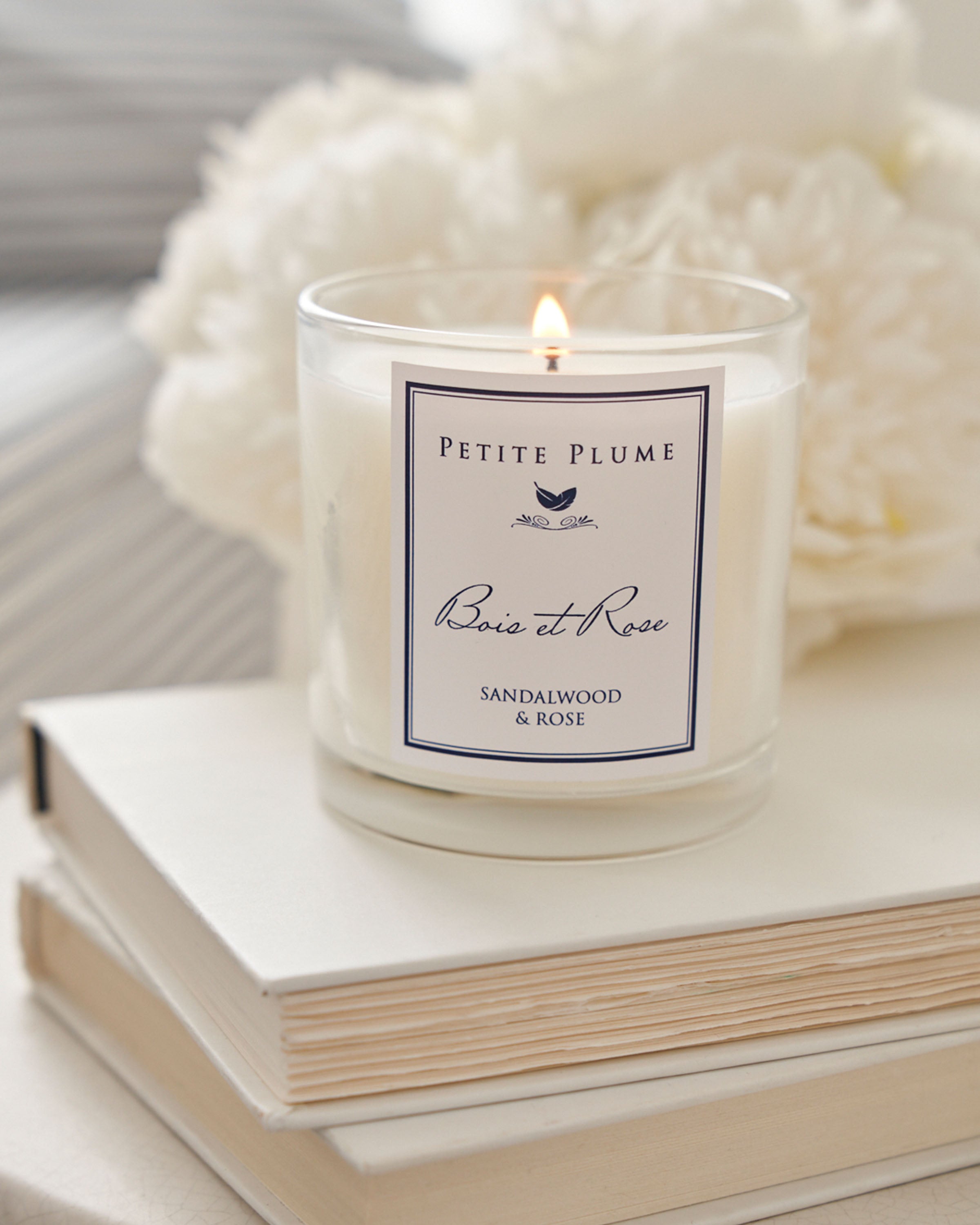 A lit Petite Plume Bois et Rose Candle from the Candle Collection sits elegantly on two stacked books, with soft-focus white flowers and striped fabric in the backdrop creating a serene ambiance.