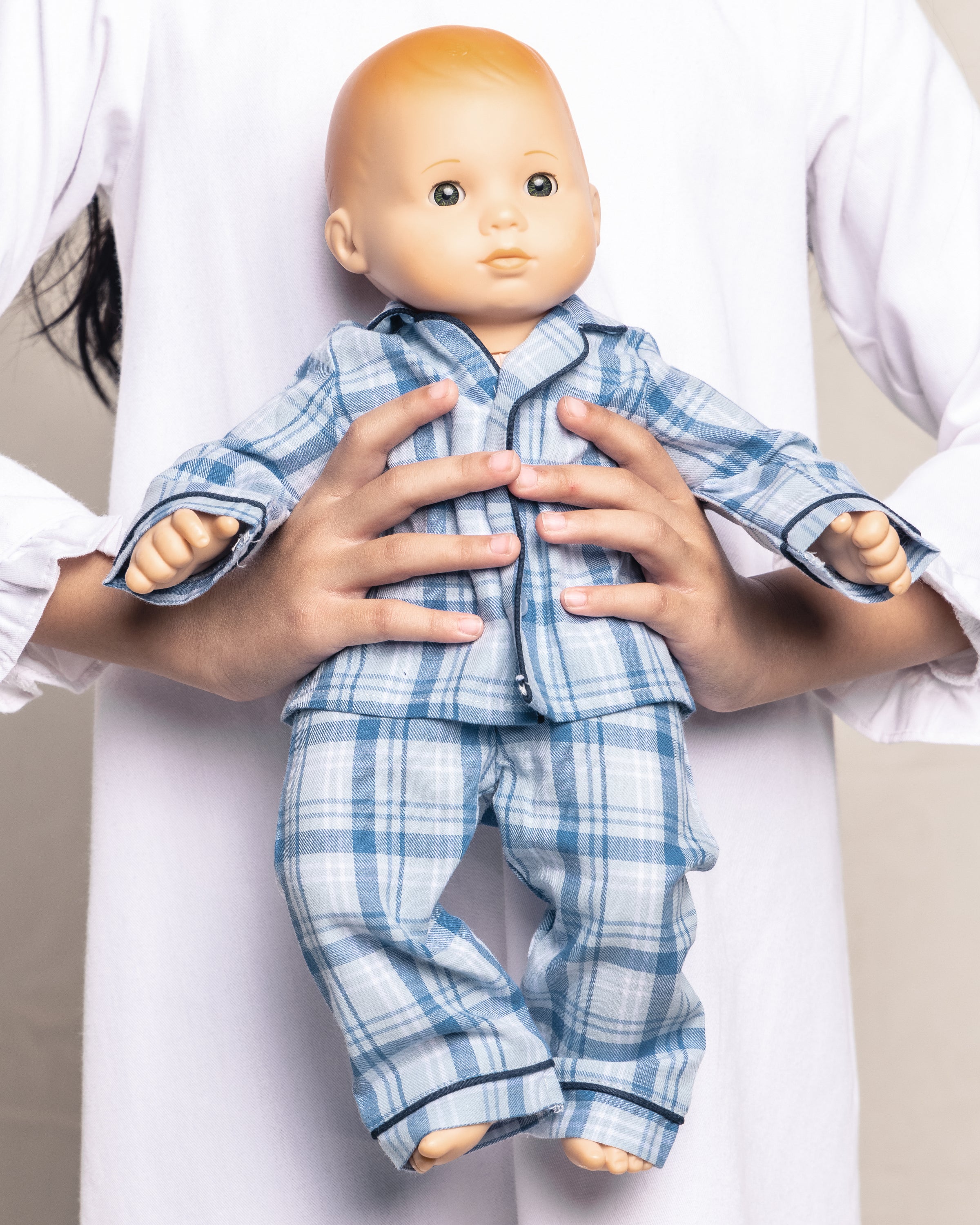 A person holds an 18 doll dressed in Petite Plumes Kids Twill Doll Pajamas in Seafarer Tartan, perfectly matching their white long-sleeve shirt against a plain, light-colored background.