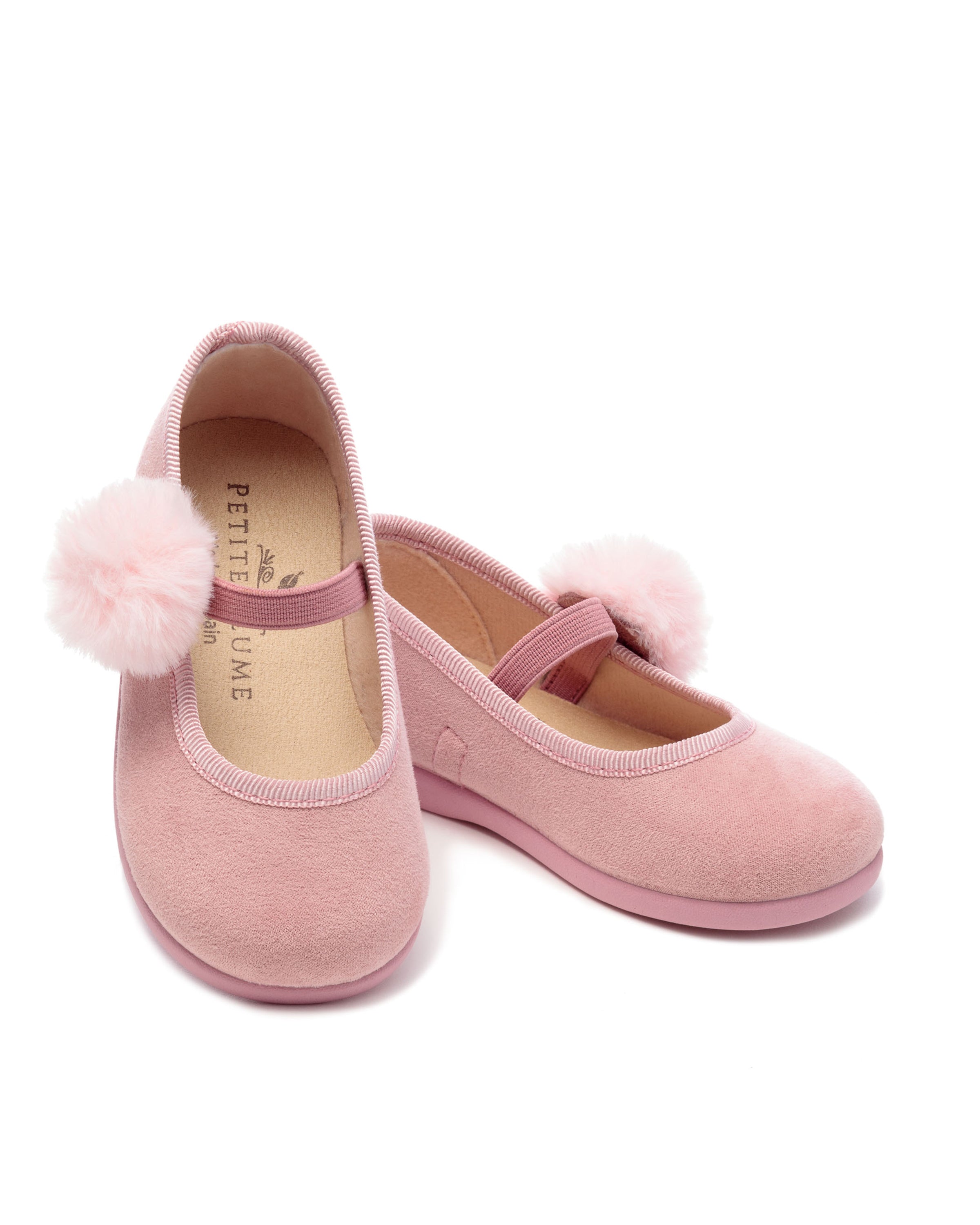 The Kids Delphine Slipper in Antique Rose Suede by Petite Plume features soft texture, elastic straps, and festive pom-poms on the toes. These artisan slippers have a tan lining, elegantly displayed on a plain white background.
