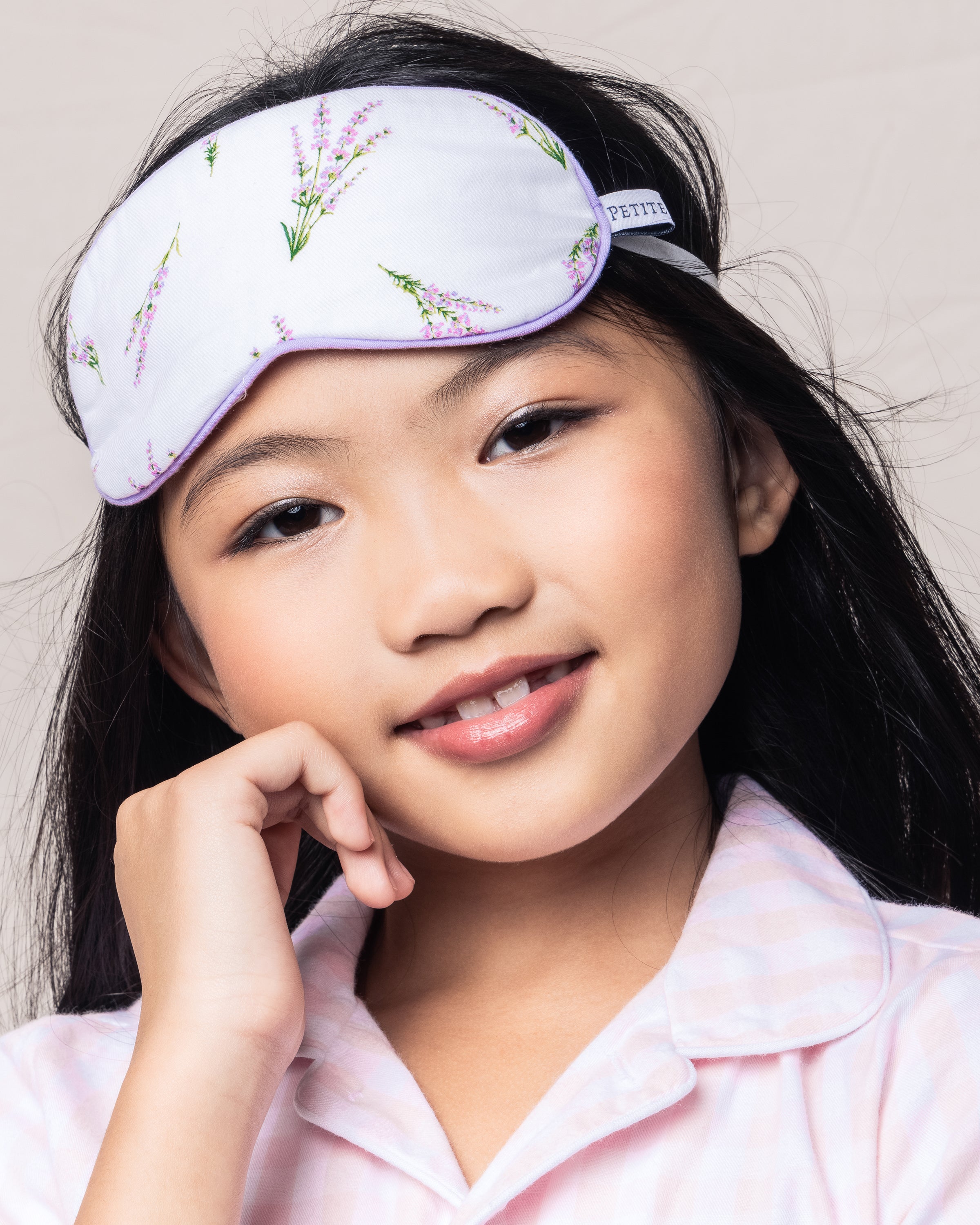A young girl with long black hair smiles softly, wearing the Kids Sleep Mask in Fields of Provence by Petite Plume. This luxury piece complements her light pink plaid pajama top as her hand gently touches her cheek.