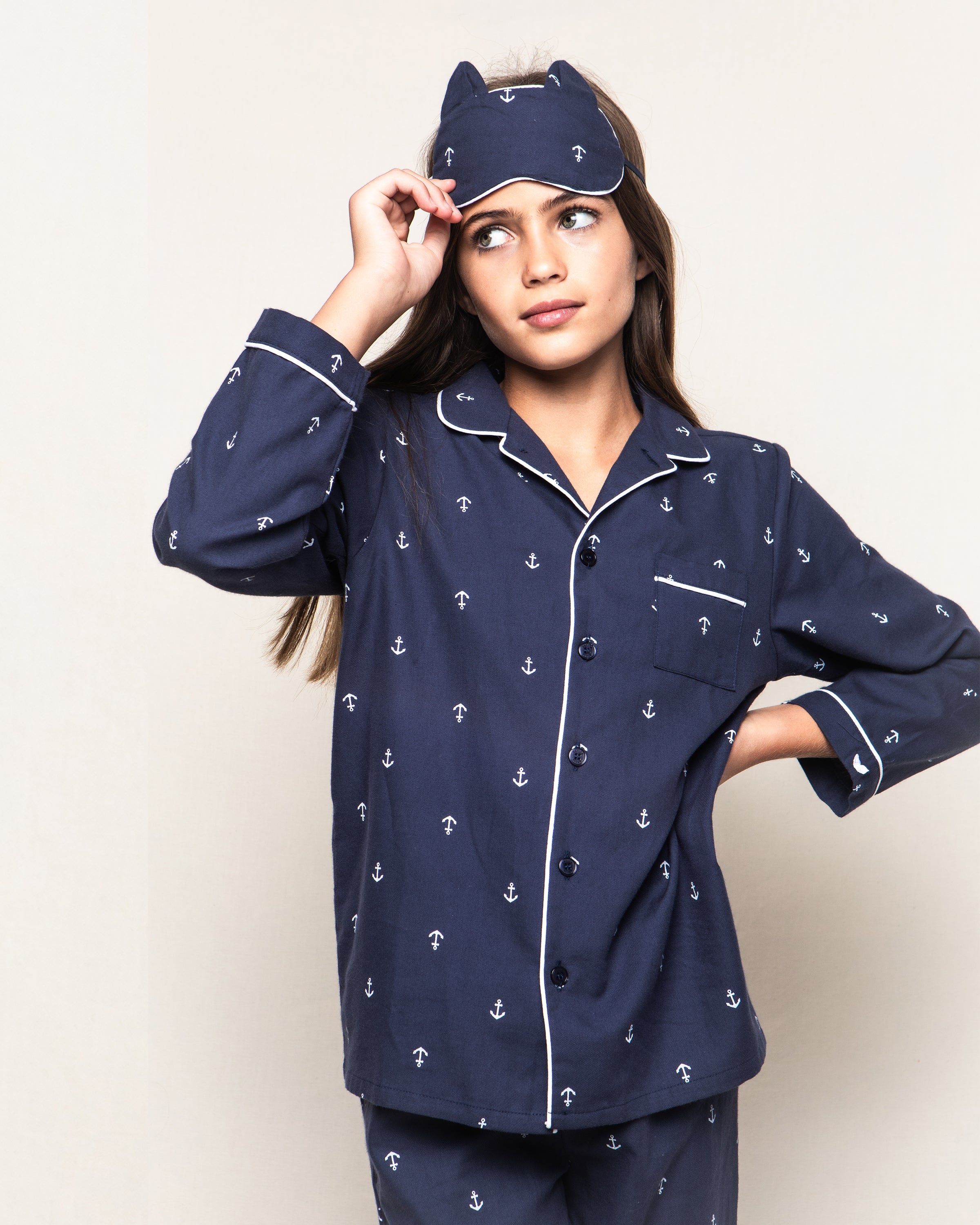 A girl in Petite Plumes navy Kids Twill Kitty Sleep Mask in Portsmouth Anchors adjusts it, gazing sideways against a plain backdrop. This elegant sleepwear set with a matching eye mask radiates comfort and charm, ideal for stylish bedtime relaxation.