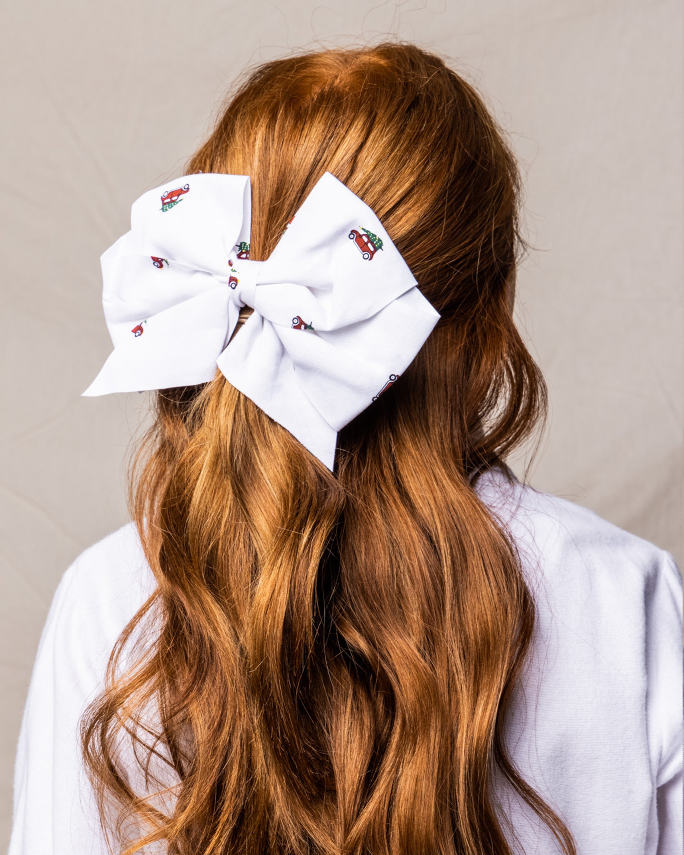 A person with long red hair models the Petite Plume Girls Hair Bows in Holiday Journey, a festive white bow featuring red and black ladybug patterns, ideal as a holiday gift. The scene is set against a soft, neutral background.
