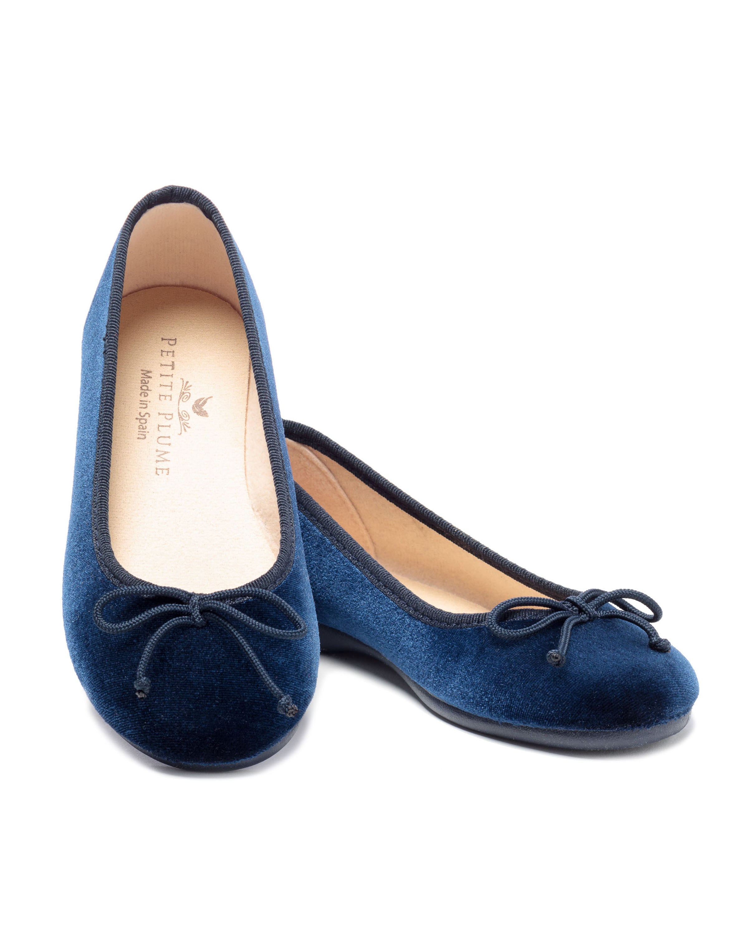 The Kids Juliette Ballet Slipper in Navy Velvet from Petite Plume, featuring bows on the toes and light-colored branded insoles, exude a luxe suede allure. They are elegantly positioned with one slightly ahead against a pristine white background.