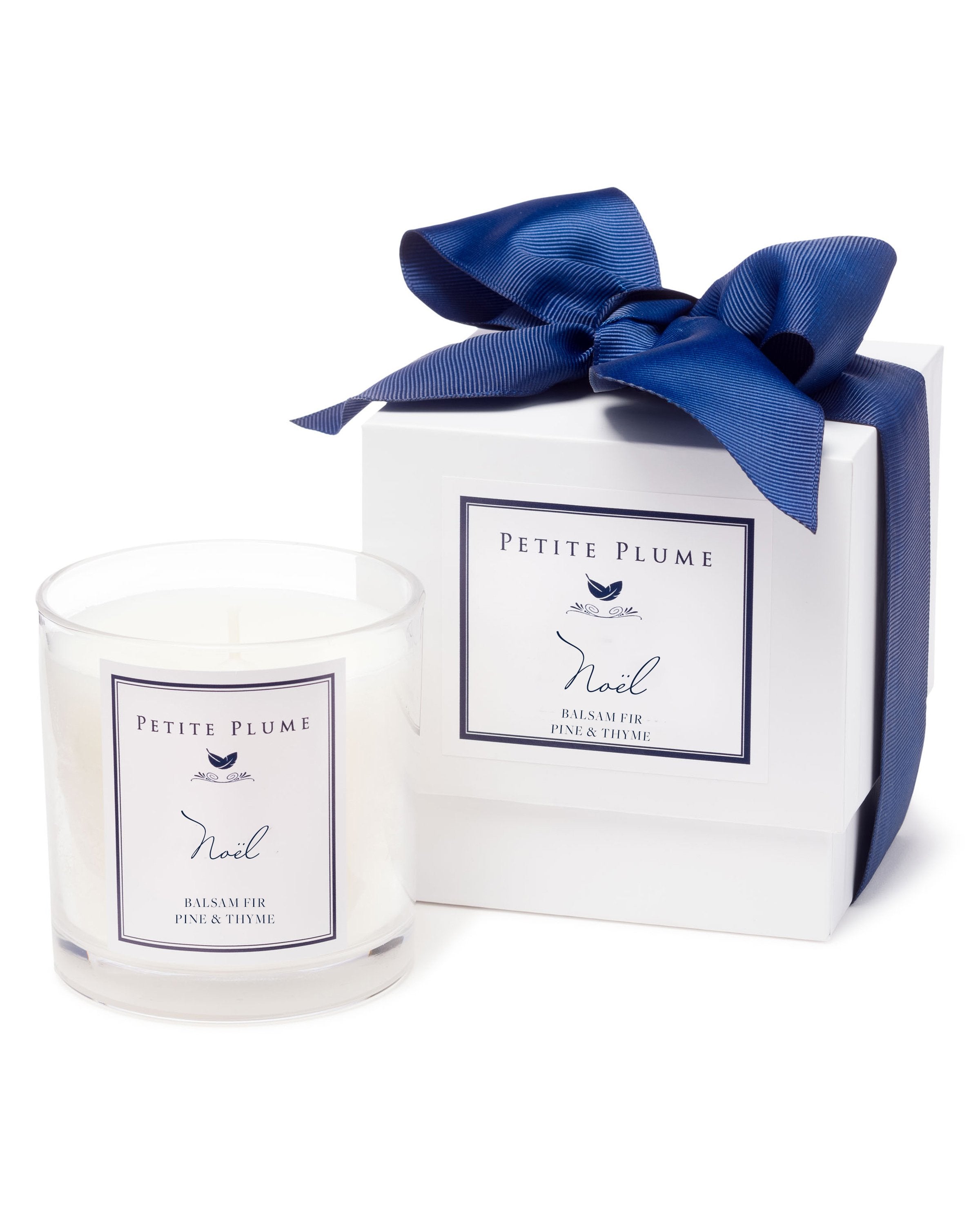 The Petite Plume Noël Candle, featuring Balsam Fir, Pine & Thyme scents, sits in a glass holder beside an elegant white box with a blue ribbon from their Candle Collection.