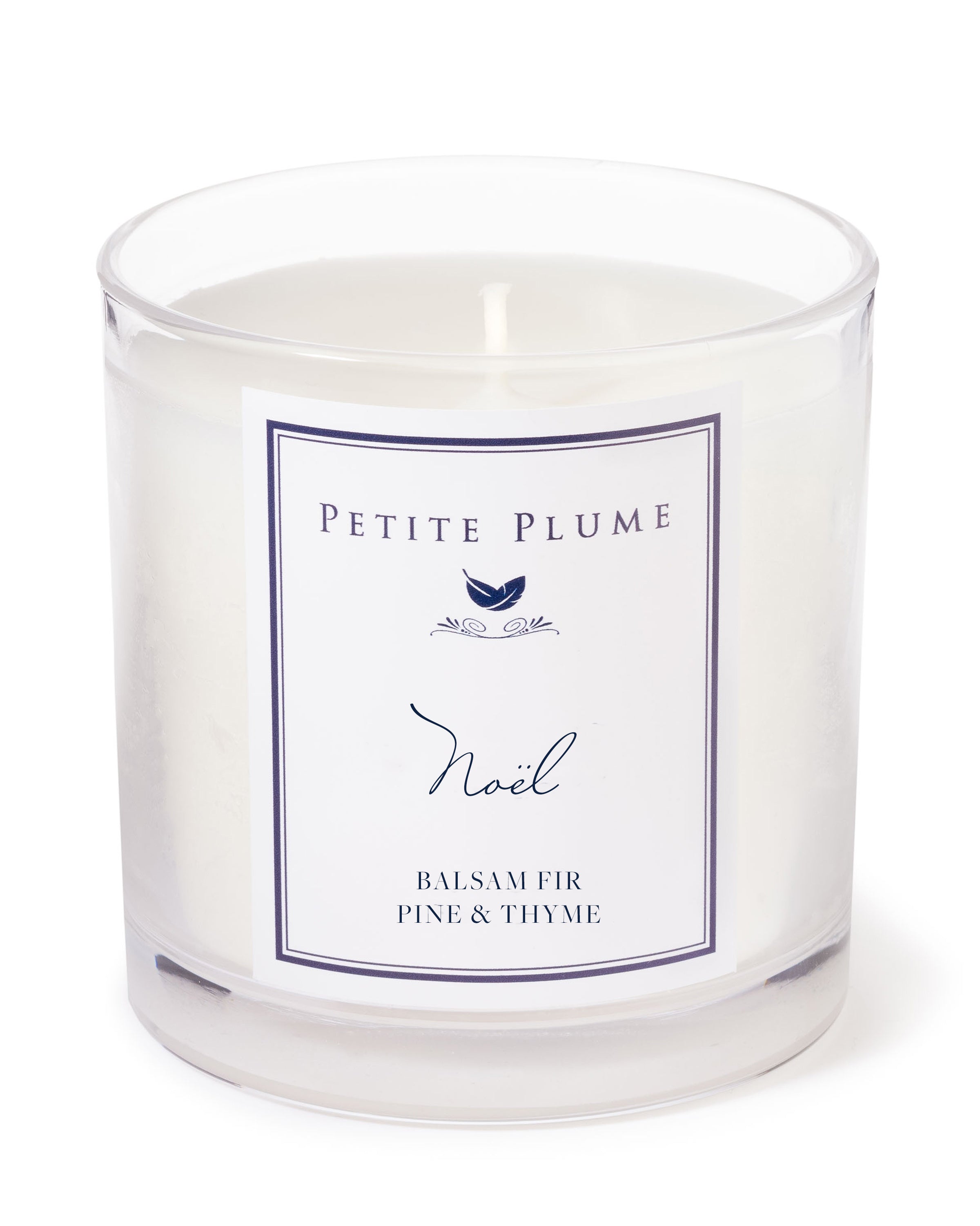 The Petite Plume Noël Candle from the Petite Plume Candle Collection is a clear glass candle with a white single wick, scented with balsam fir, pine, and thyme. Its label features a simple design with a small bird logo.