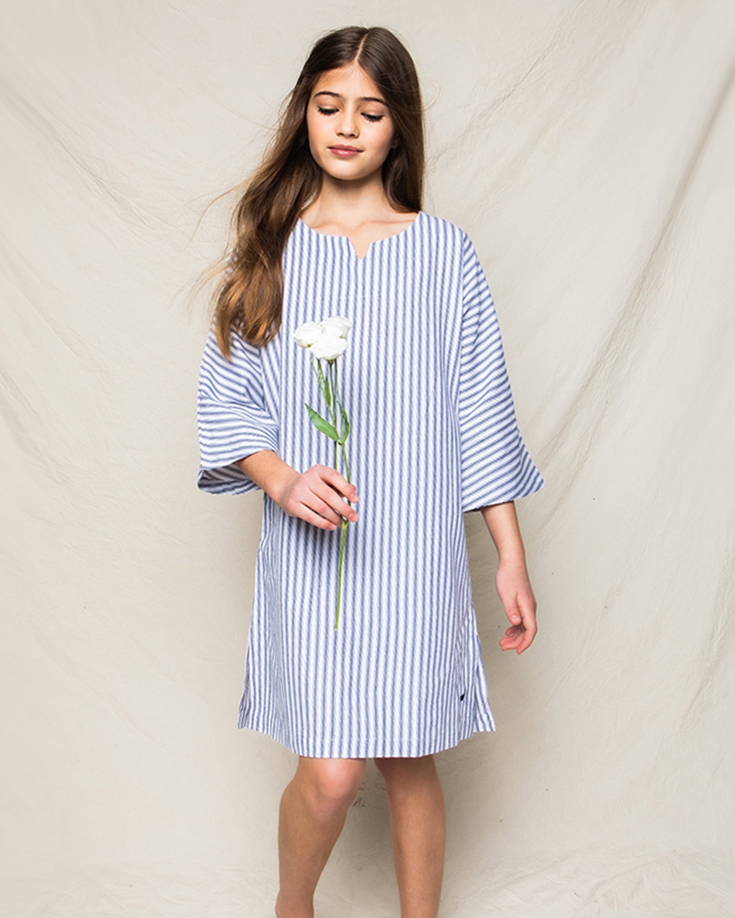 A woman in Petite Plumes Girls Twill Caftan in Navy French Ticking stands against a plain backdrop, holding a white flower. Her long hair drapes over her shoulders as she looks down slightly.