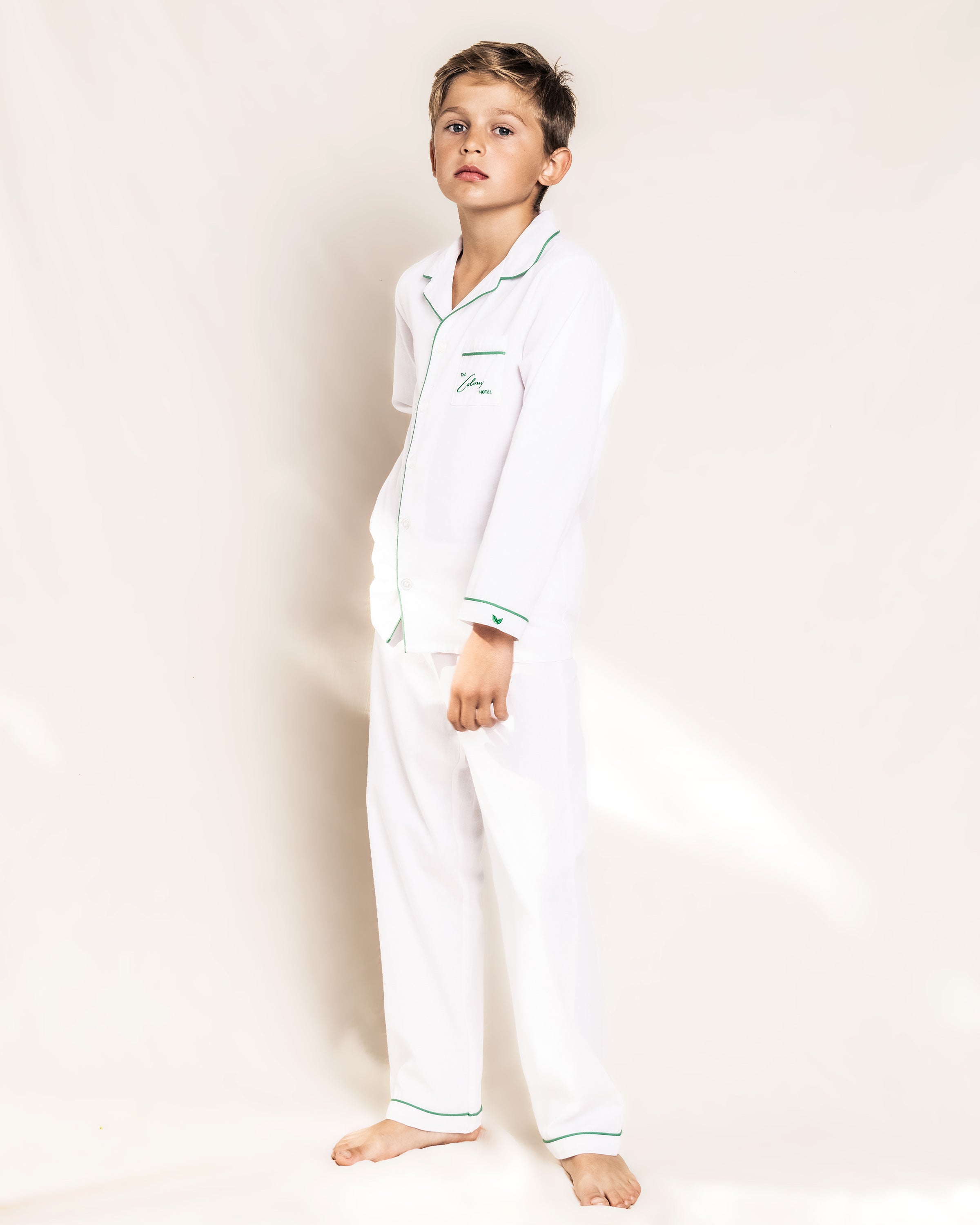 A young boy stands barefoot wearing the Colony Hotel x Petite Plume Kids Pajama Set, featuring white fabric with green piping. He looks slightly to the side with a neutral expression, embodying comfort and elegance similar to a stay at the Colony Hotel.