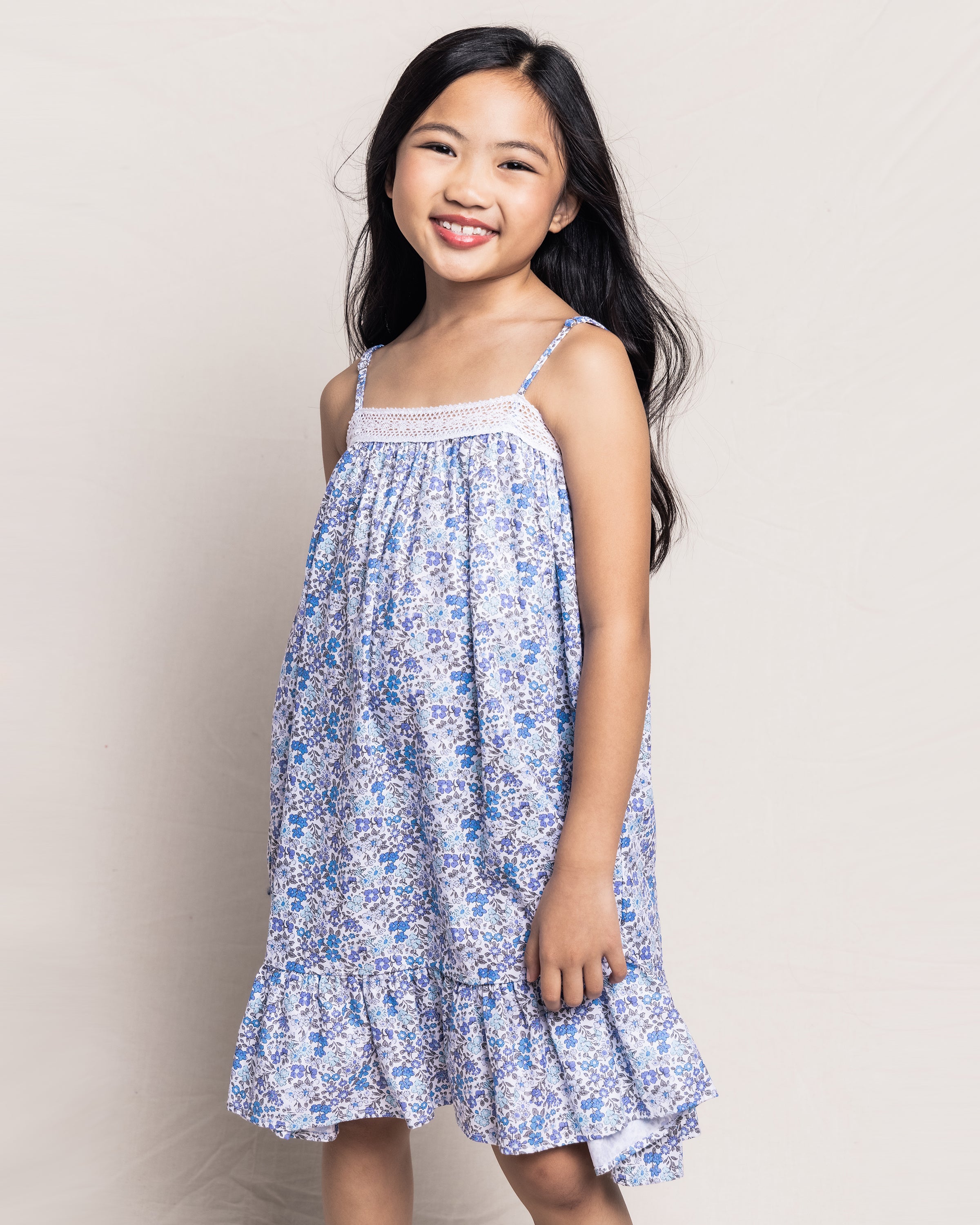 A young girl with long black hair smiles, wearing Petite Plumes Girls Twill Lily Nightgown in Fleur DAzur, featuring a vintage-inspired floral print and a ruffled hem. She stands against a plain, light-colored background.