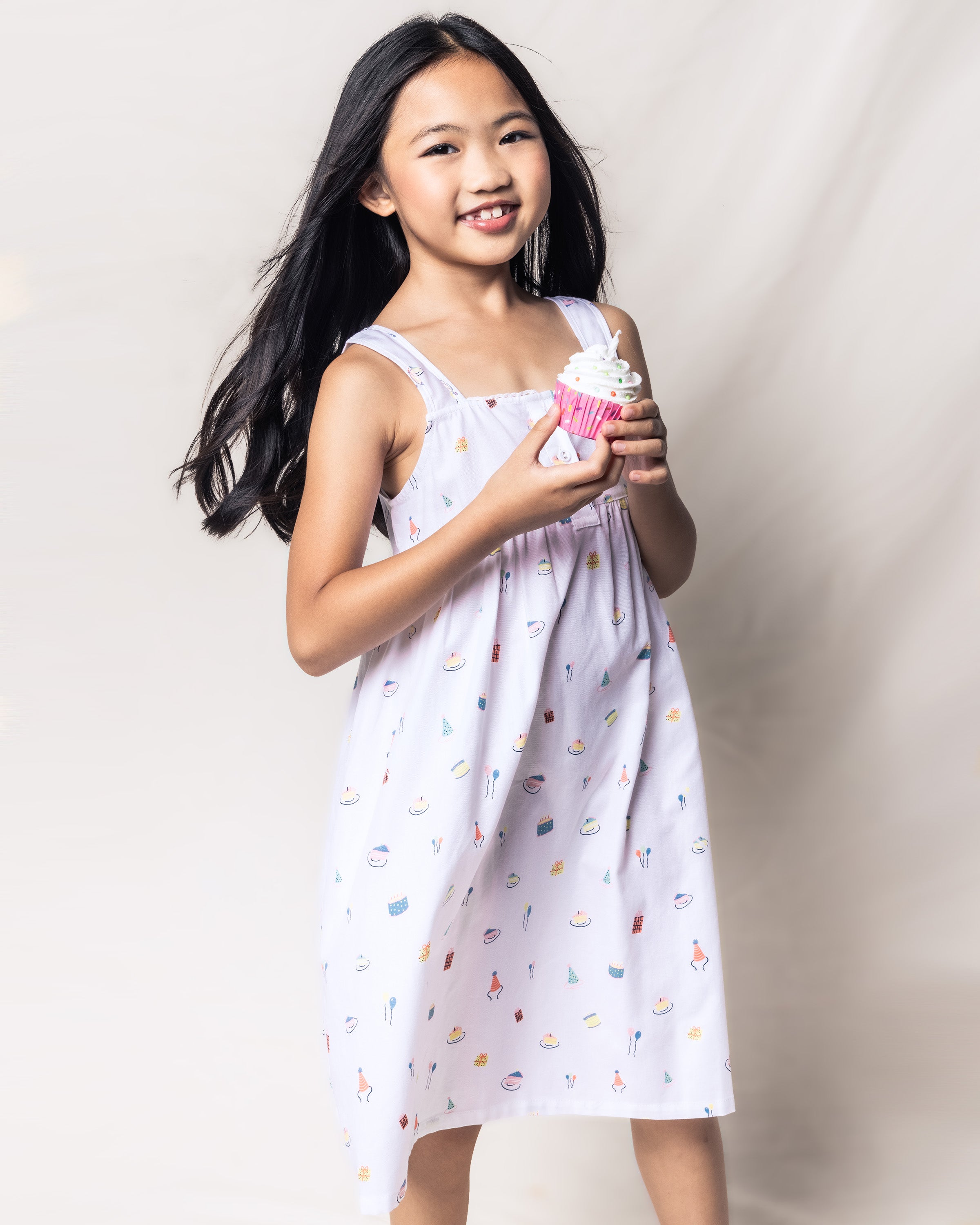 A young girl smiles while holding a cupcake with white frosting. She wears the Petite Plume Girls Twill Charlotte Nightgown in Birthday Wishes, crafted from yarn-dyed cotton with colorful patterns, and stands against a light, neutral background. Her long black hair cascades over her shoulders.