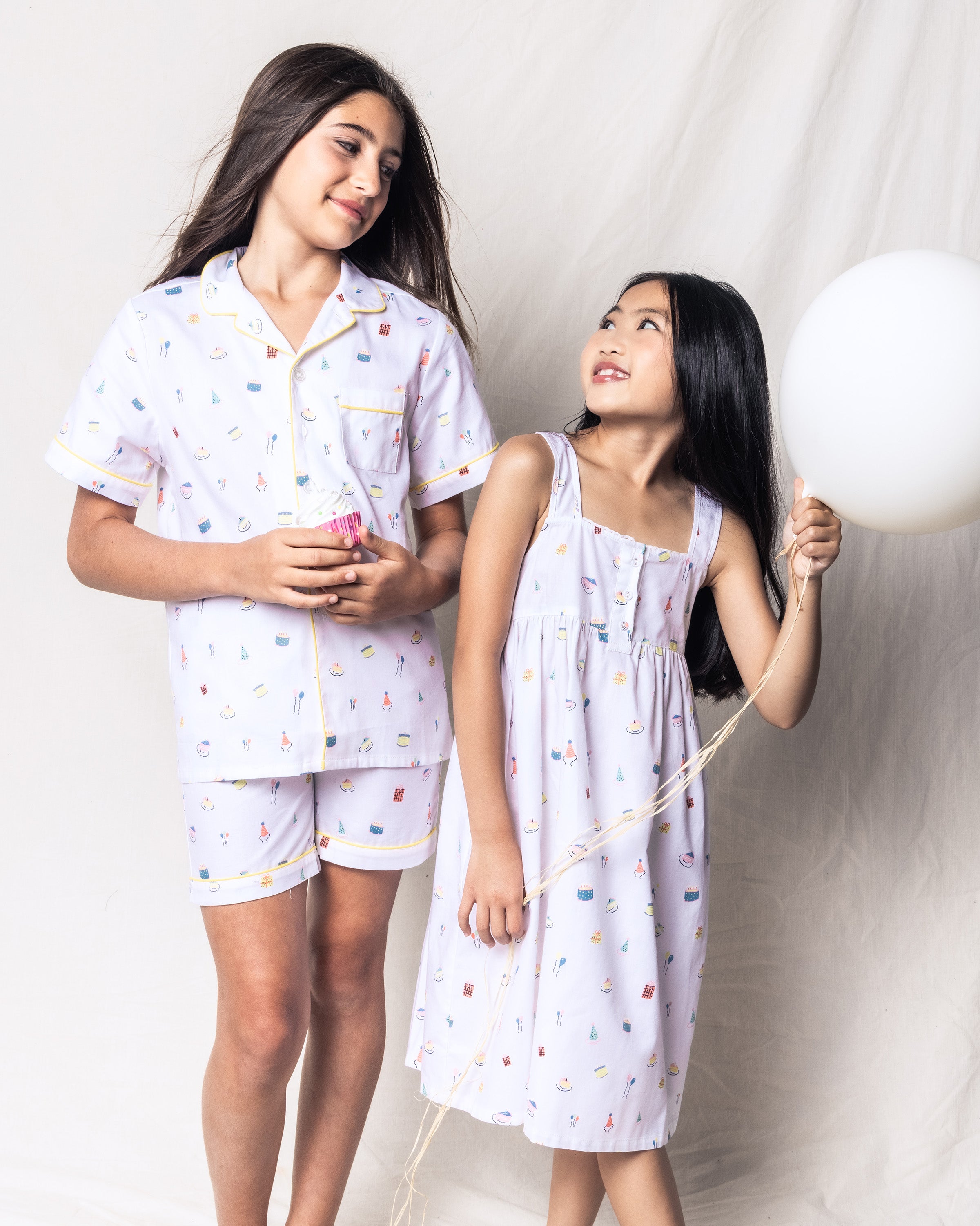 Two young children smile at each other, wearing Petite Plumes matching Girls Twill Charlotte Nightgown in Birthday Wishes. One child holds a cupcake, and the other grips a white balloon against a light, neutral background, radiating joy in their cozy sleepwear.