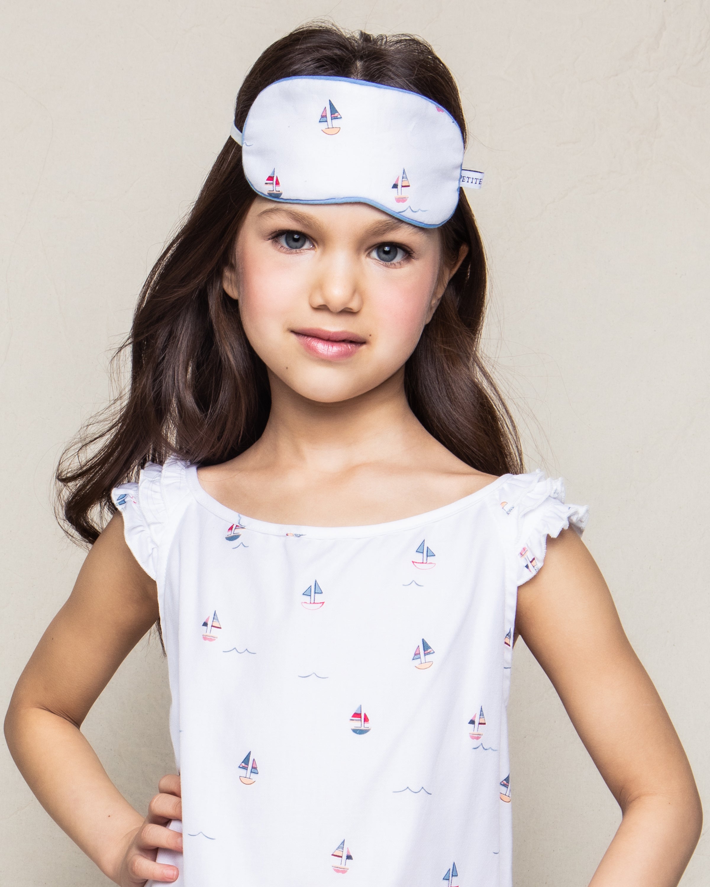 Against a plain background, a young girl exudes charm, wearing Petite Plumes Kids Twill Sleep Mask in Bateau, adorned with sailboats to match her sailboat-patterned dress. She stands with a slight smile and hands on her hips, showcasing this monogrammed delight.