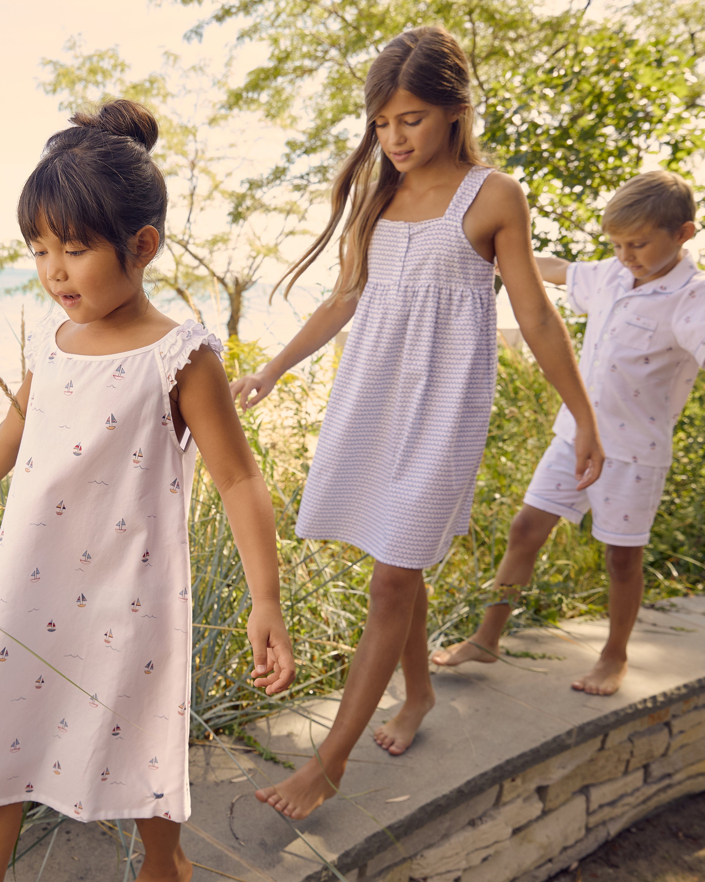 Three barefoot children stroll on a stone path near the beach. The leading girl wears a Petite Plume Girls Twill Charlotte Nightgown in La Mer. The middle girl dons a light blue dress, and the boy at the back is in a white shirt and shorts, with trees and sand creating a serene backdrop.