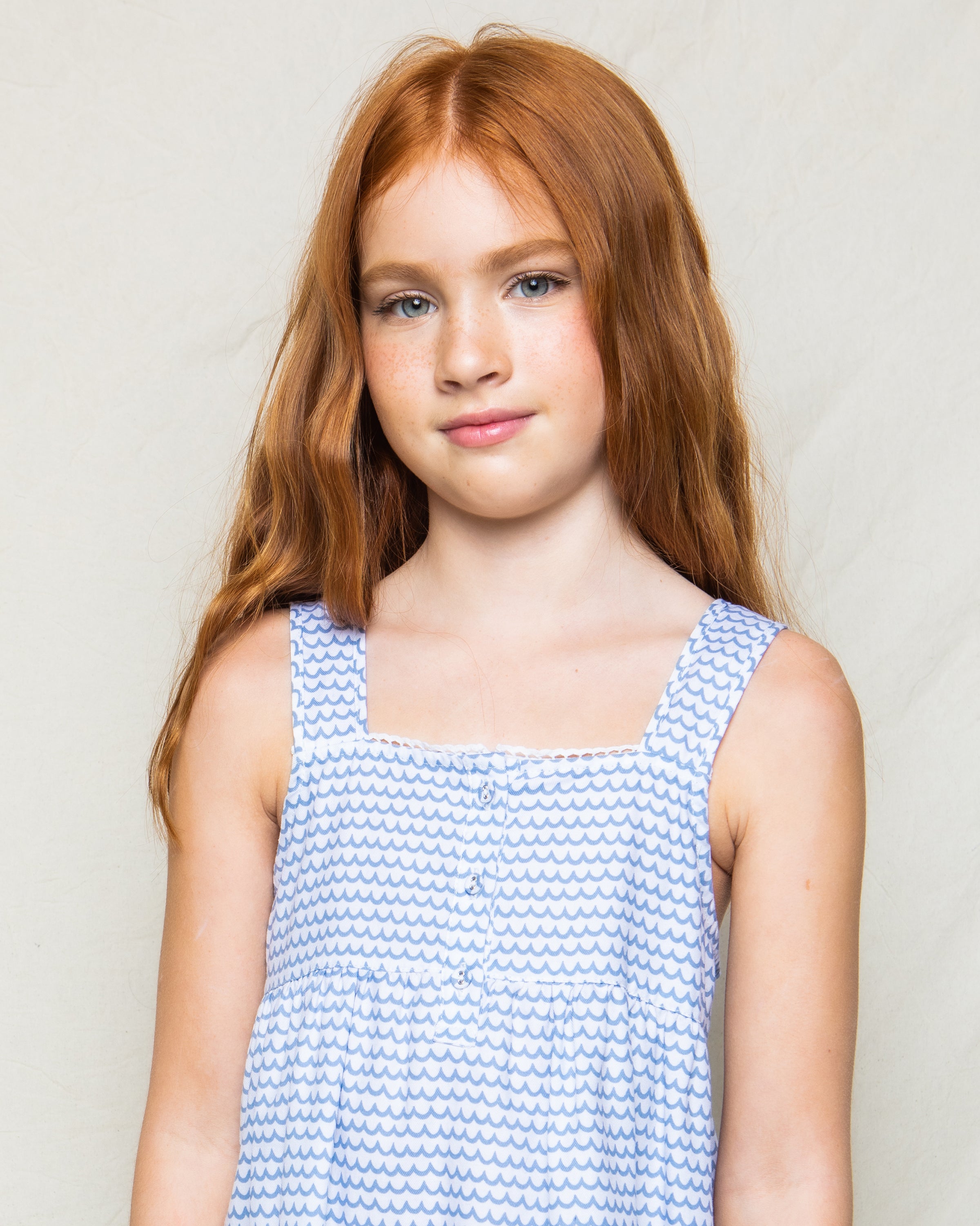 A young girl with long red hair stands gracefully in a charming Girls Twill Charlotte Nightgown in La Mer by Petite Plume, the white and blue patterns softly complimenting the plain background.