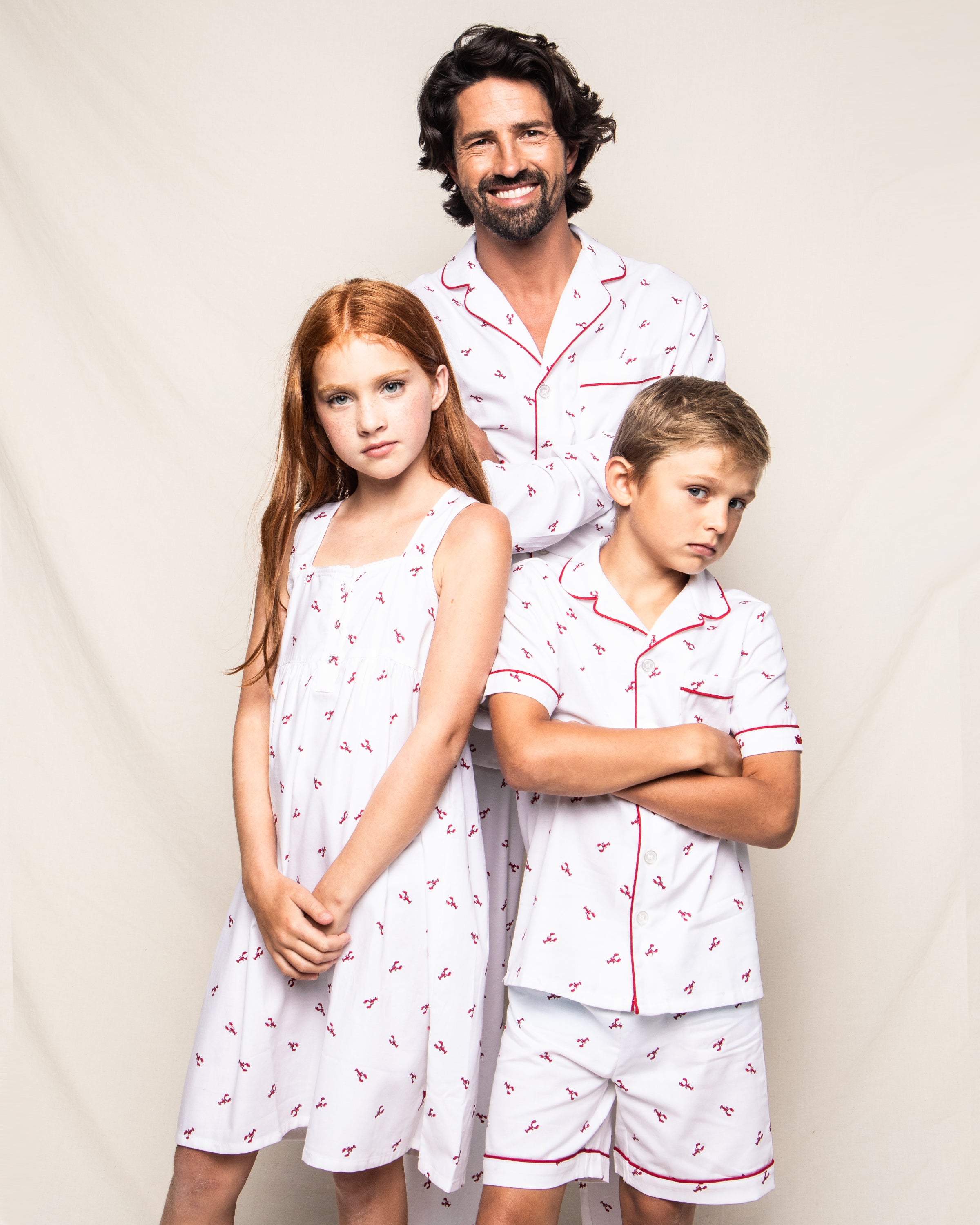 A man and two children pose against a plain background, all wearing Petite Plumes Brixham Lobsters sleepwear. The girl sports a nightgown while the boy and man don Kids Twill Pajama Sets. Their red hair complements the luxurious sleepwear with red piping and designs.