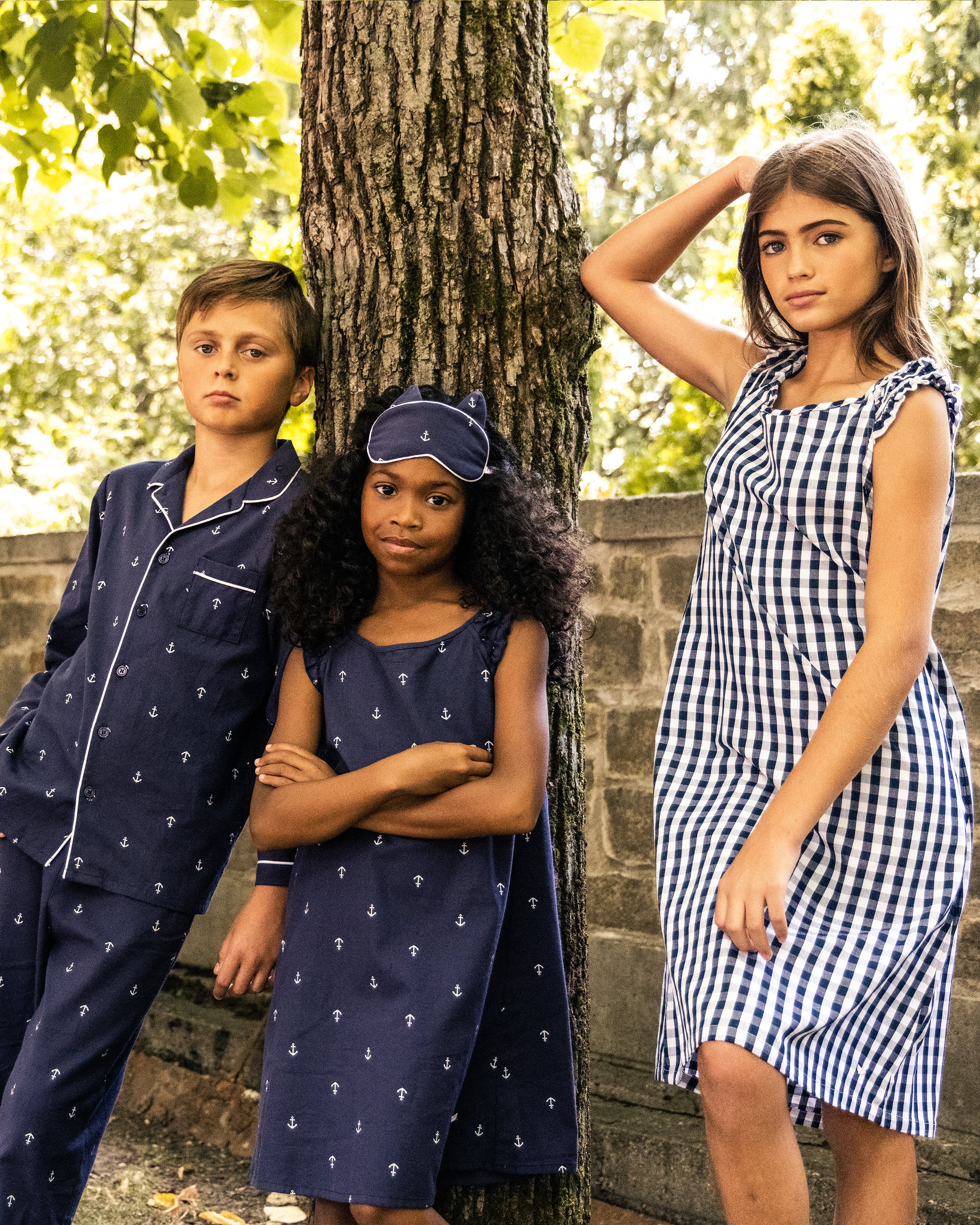 Three kids pose under a tree. A boy in Petite Plume Kid’s Twill Pajama Set in Portsmouth Anchors leans against the trunk. A girl in a matching nightgown and cap stands with hands crossed, while another girl in a blue checkered dress rests an arm on the tree, creating a serene scene of luxurious sleepwear bliss.