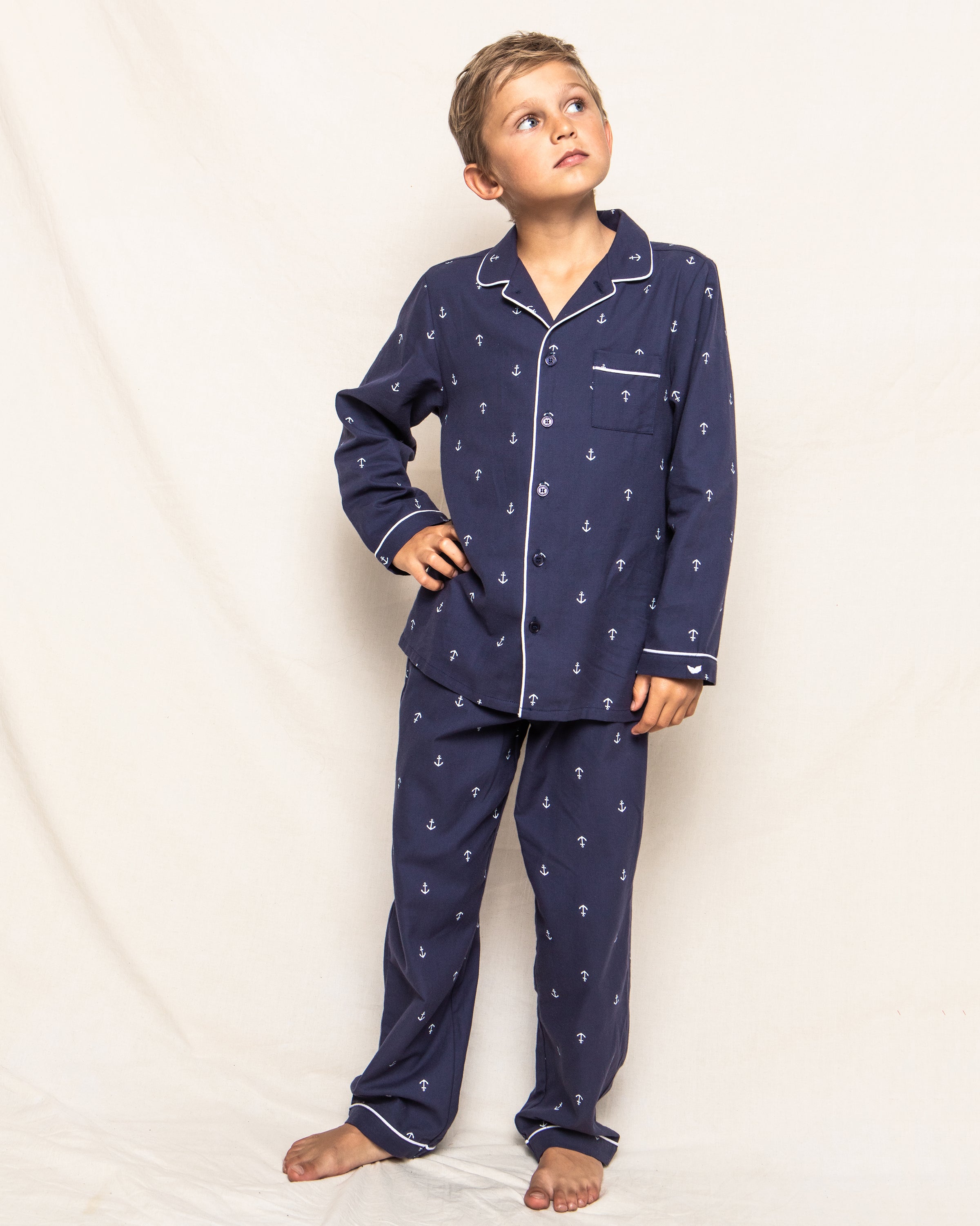 A young boy stands barefoot against a plain background, looking upward. Wearing Petite Plumes Kids Twill Pajama Set in Portsmouth Anchors, featuring a navy blue anchor pattern with white trim, his hand rests on his hip. He embodies both comfort and style in this luxury sleepwear.