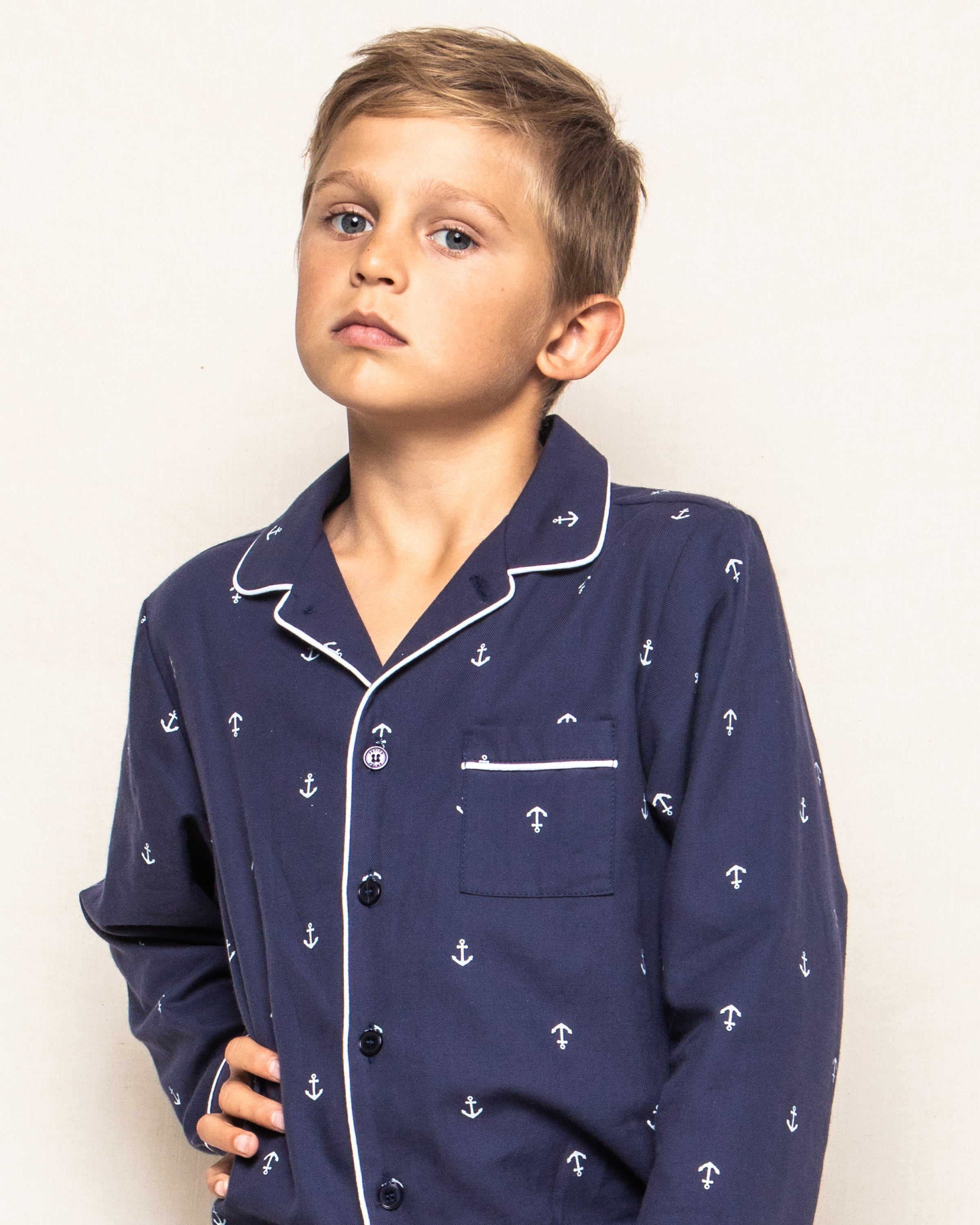 A young boy with short hair wears the Kids Twill Pajama Set in Portsmouth Anchors by Petite Plume, showcasing his love for nautical pajamas. He stands with a hand on his hip, looking slightly to the side with a serious expression against a plain, neutral background.
