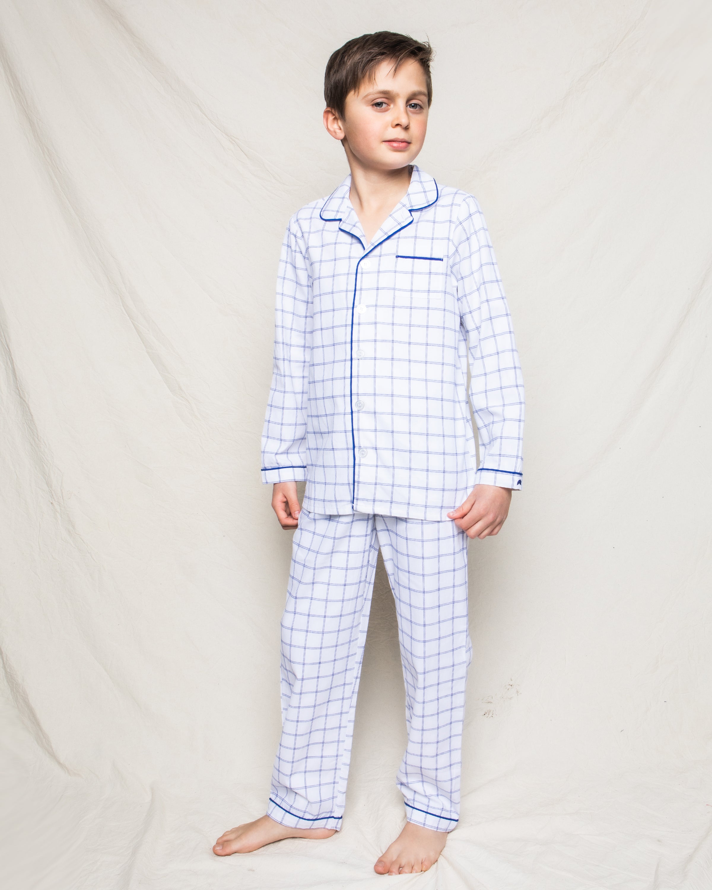 A young boy with short dark hair stands barefoot on a plain background, wearing Petite Plumes Kids Twill Pajama Set in Nantucket Tattersall. His expression is neutral, and he gazes slightly to the side.