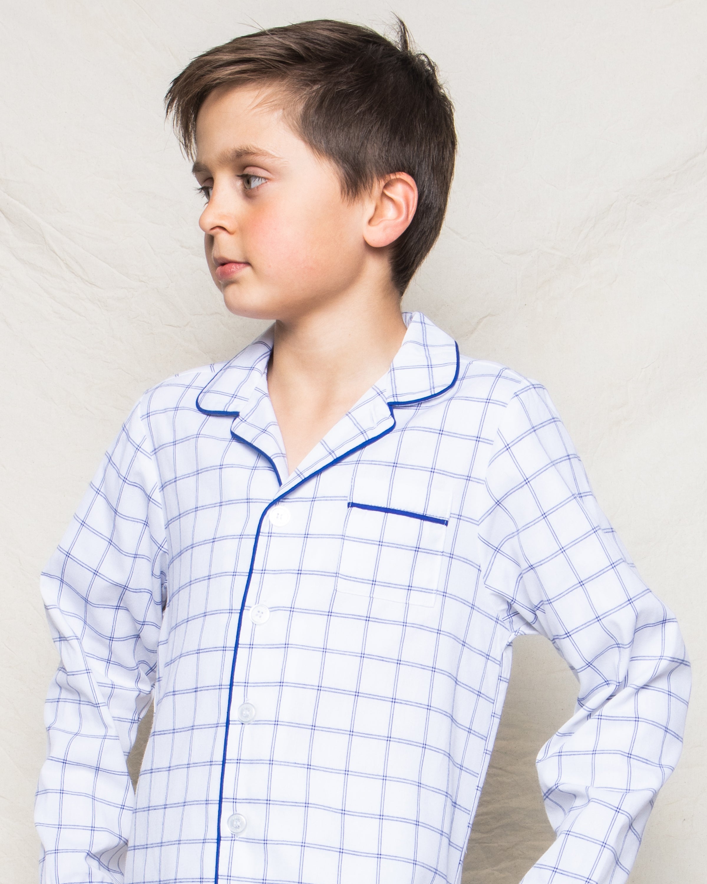A young boy wears the Kids Twill Pajama Set in Nantucket Tattersall by Petite Plume, showcasing a white checkered pattern with blue piping. He poses against a neutral background, gazing to the side with a calm expression.
