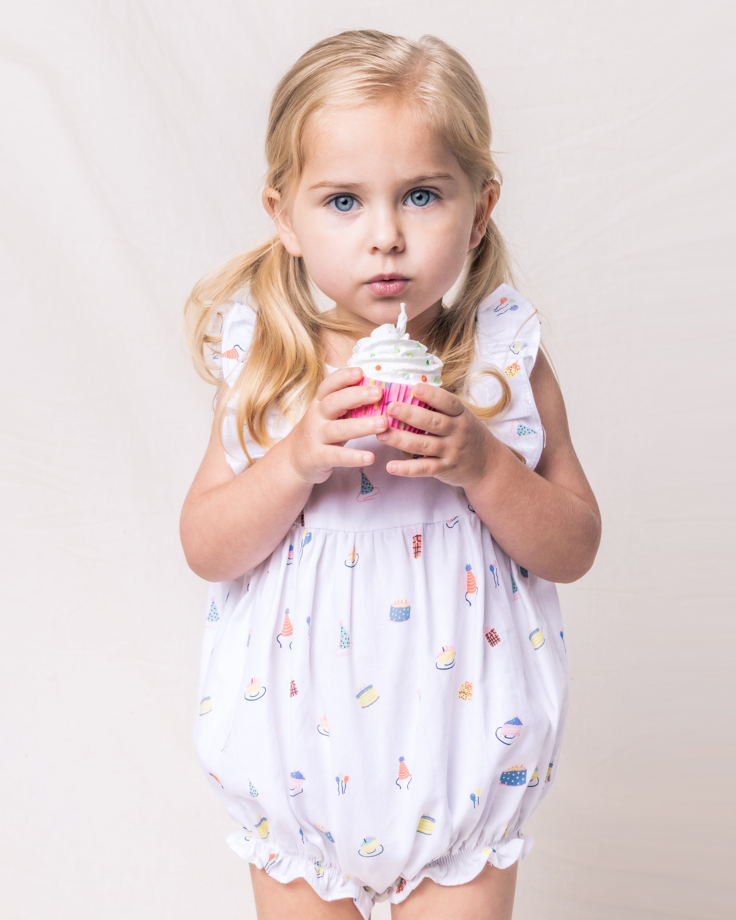 A blonde child with pigtails holds a cupcake with white frosting and a candle. She wears Petite Plumes Babys Twill Ruffled Romper in Birthday Wishes, made from yarn-dyed cotton, and displays a curious expression against a plain, light background.