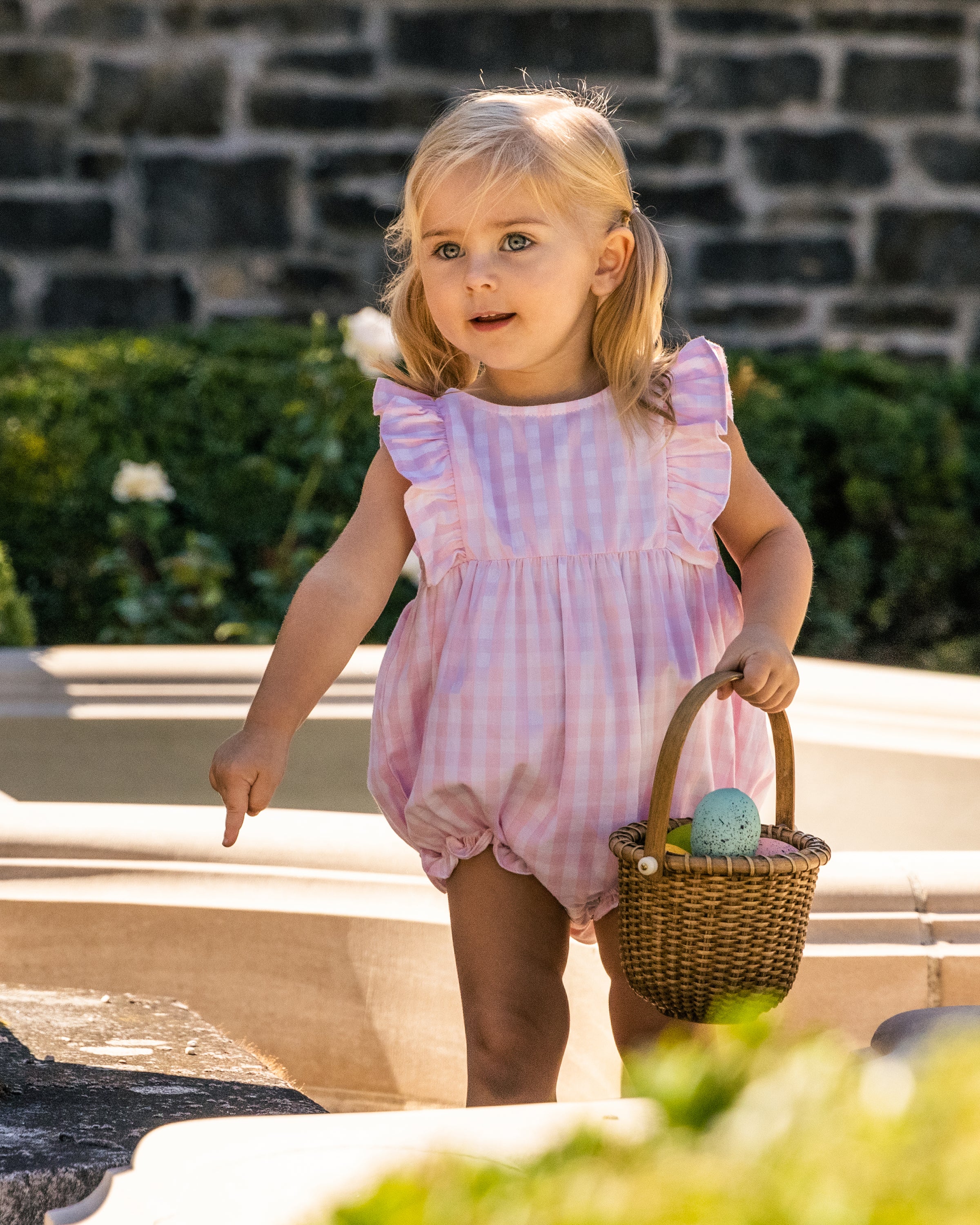 A young child with blonde hair is wearing Petite Plumes Babys Twill Ruffled Romper in Pink Gingham. She holds a wicker basket with an Easter egg, standing outdoors before a stone wall and greenery, pointing at something with her right hand.