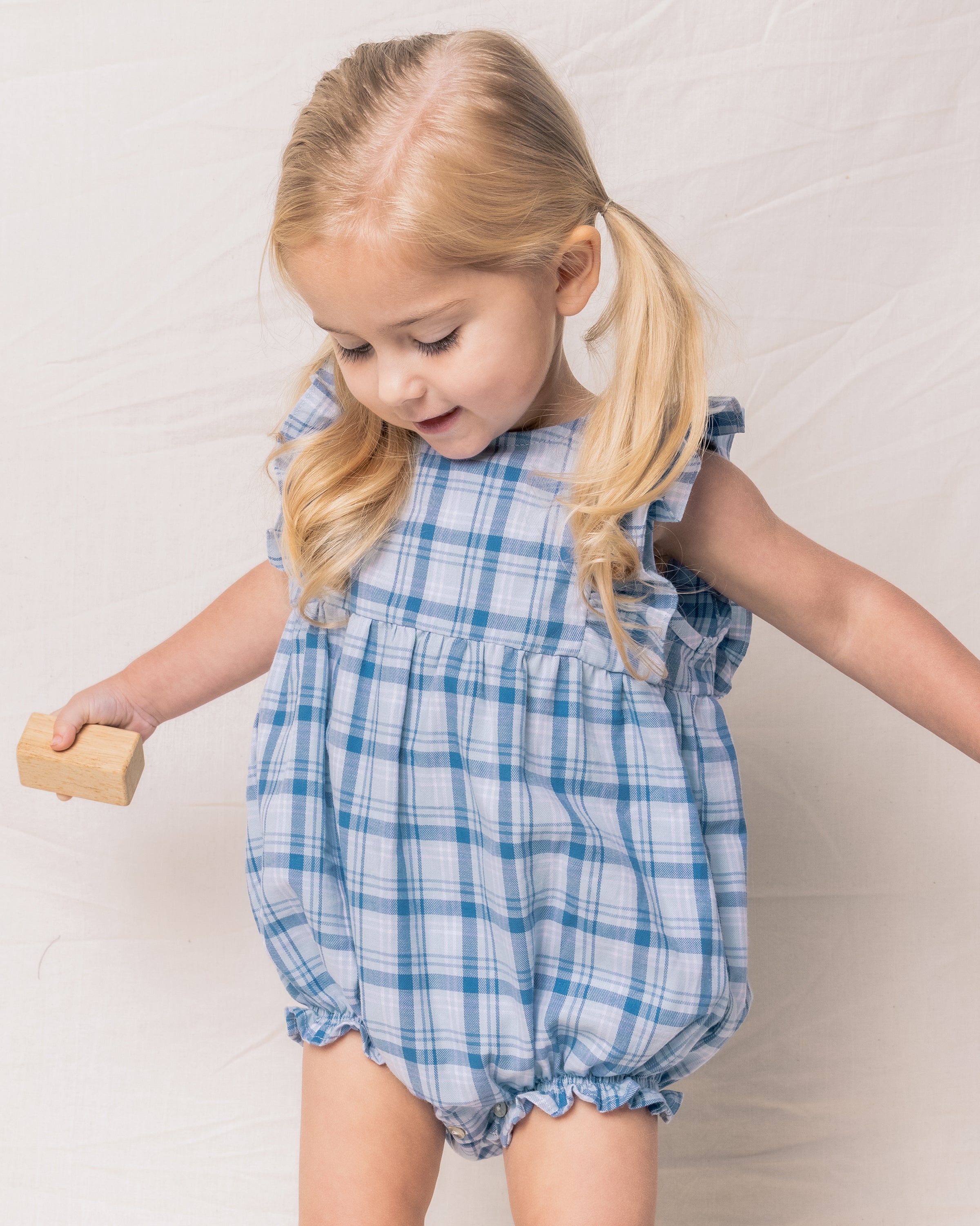 A young blonde child with pigtails is wearing Petite Plumes Babys Twill Ruffled Romper in Seafarer Tartan. Theyre holding a wooden block with a smile against a plain white background.