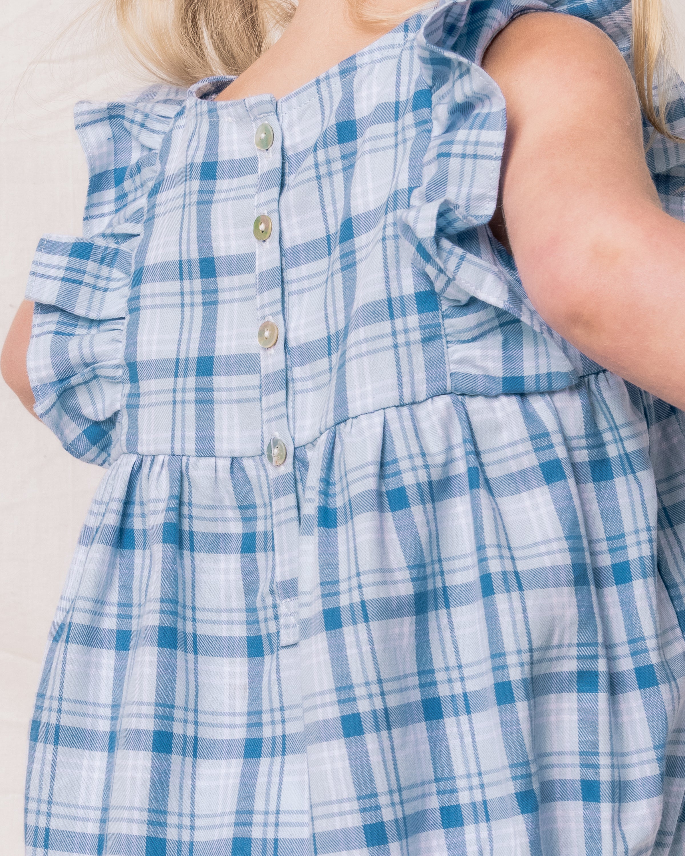 A young child wears Petite Plumes Babys Twill Ruffled Romper in Seafarer Tartan with ruffled sleeves. Buttons adorn the back of the yarn-dyed cotton romper, partially revealing the childs blond hair.