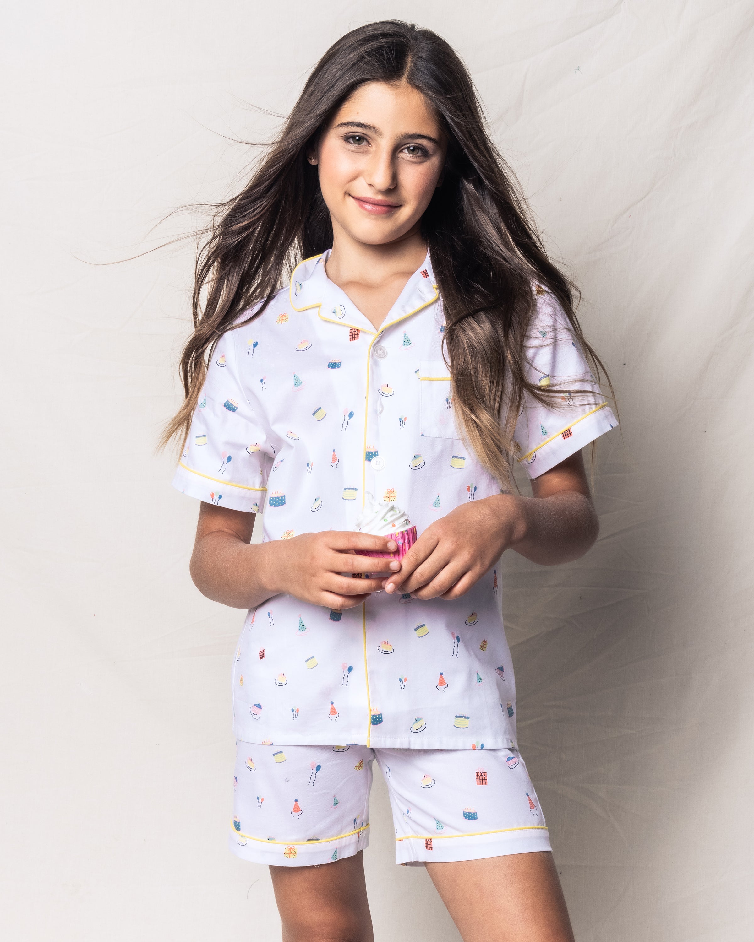 A girl with long brown hair wears Petite Plumes Kids Twill Pajama Short Set in Birthday Wishes, holding a cupcake and smiling against a light background.