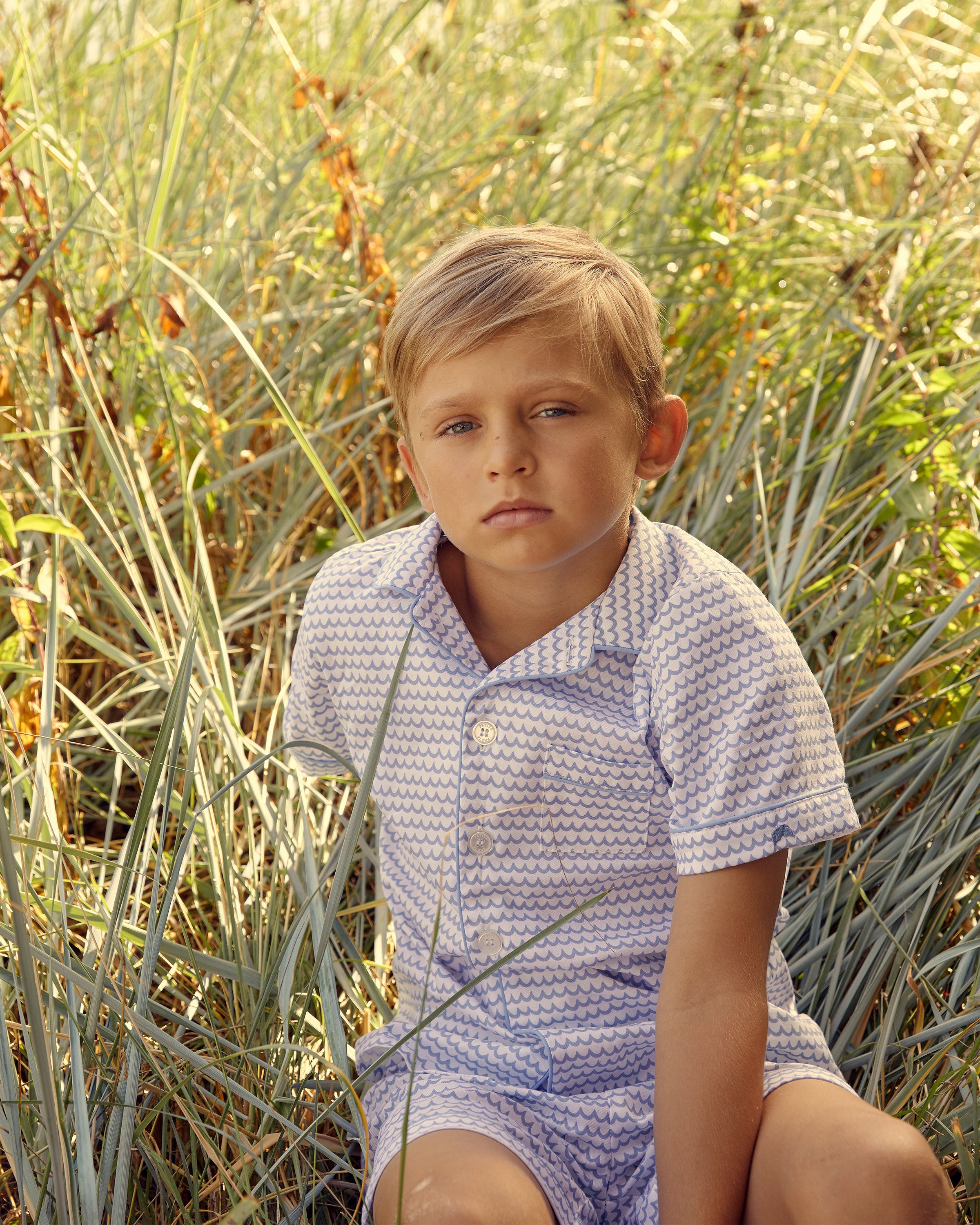 A young boy with short blond hair sits in tall grass, wearing Petite Plumes Kids Twill Pajama Short Set in La Mer. Made from the finest cotton, he gazes at the camera neutrally. The backdrop is vibrant with green and yellow grasses.