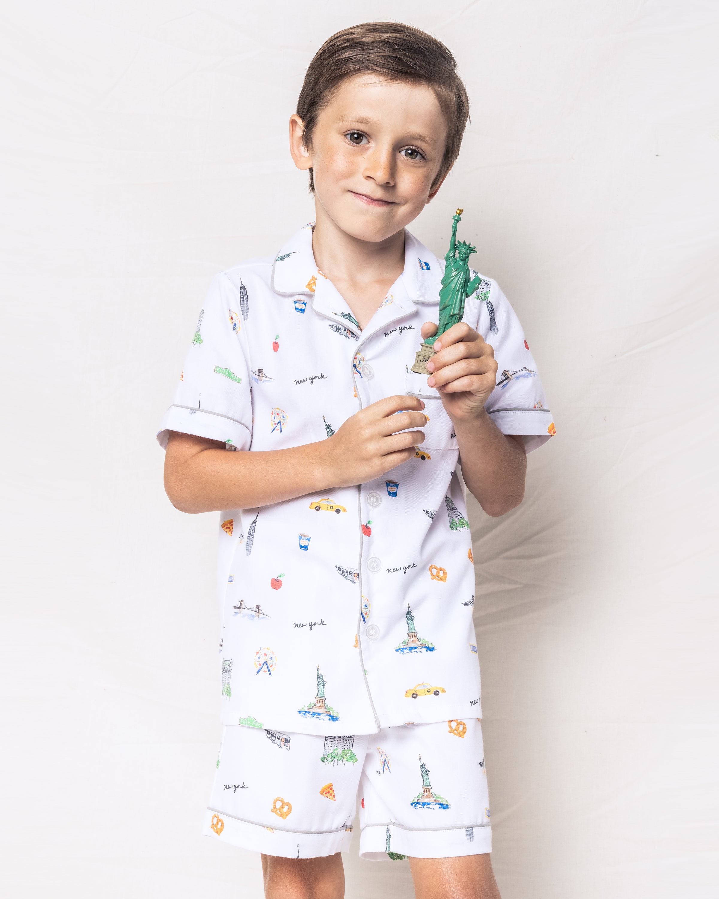 A young boy in Petite Plumes Kids Twill Pajama Short Set, featuring New York-themed illustrations, holds a small Statue of Liberty. He smiles against a backdrop of classic yellow taxis and the Brooklyn Bridge.