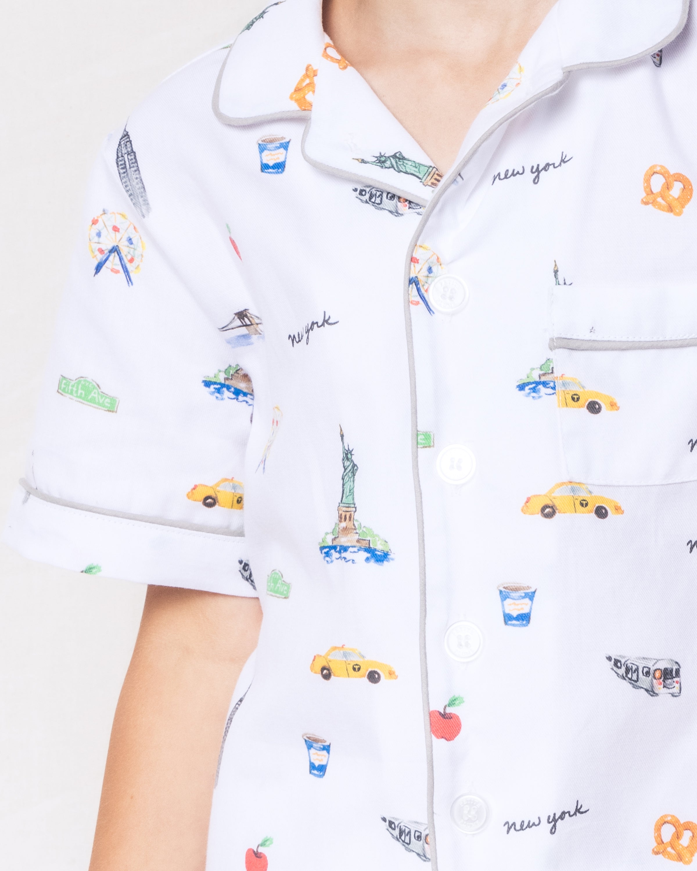 A child wears Petite Plumes Kids Twill Pajama Short Set in New York! New York! featuring illustrations of the Statue of Liberty, yellow cabs, and pretzels. Made from luxury cotton, it includes a collared shirt with gray piping for premium comfort and style.