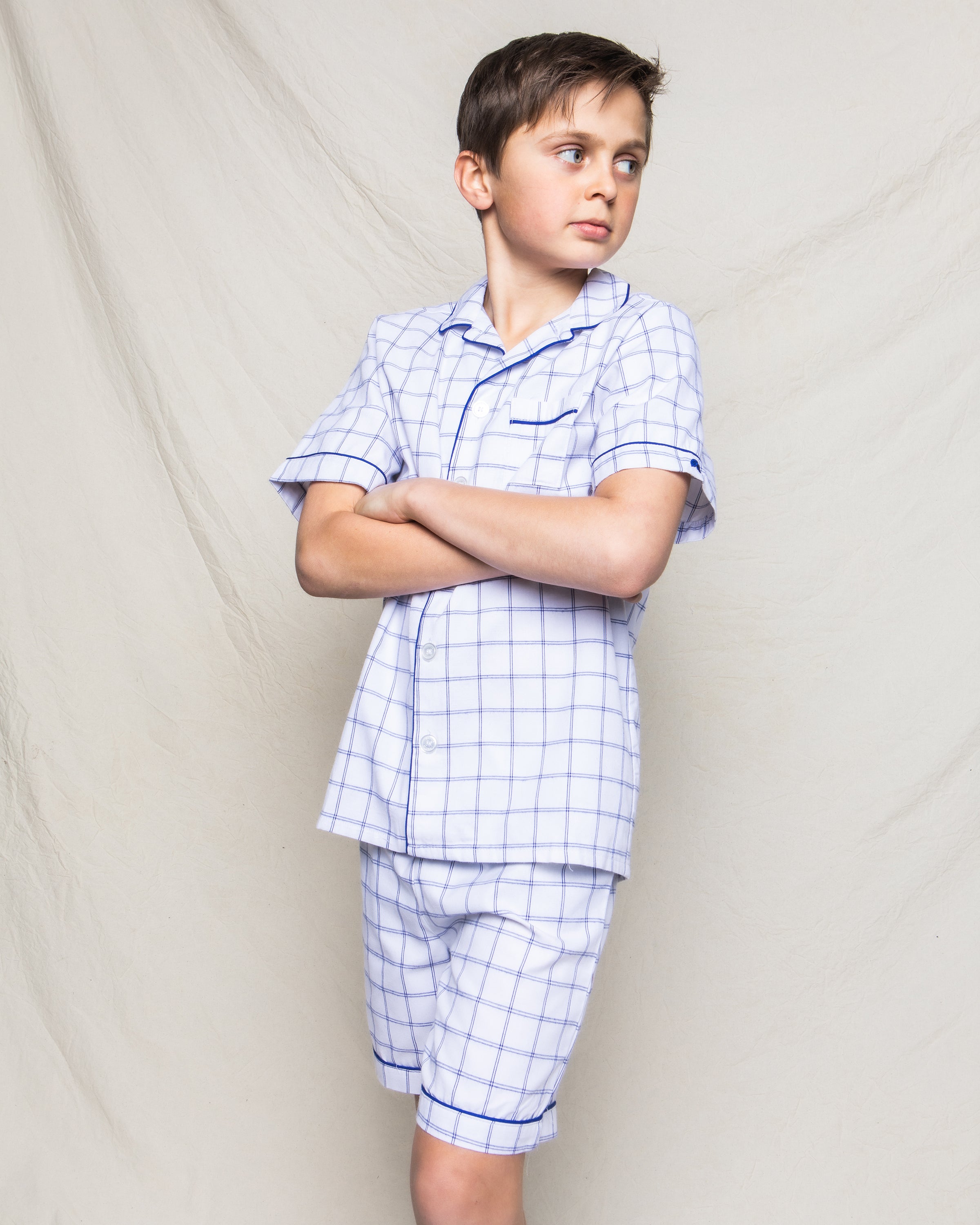 A boy wearing Petite Plumes Kids Twill Pajama Short Set in Nantucket Tattersall, with a white and blue plaid pattern, stands with arms crossed, gazing to the side. The yarn-dyed cotton complements the neutral, textured fabric backdrop.