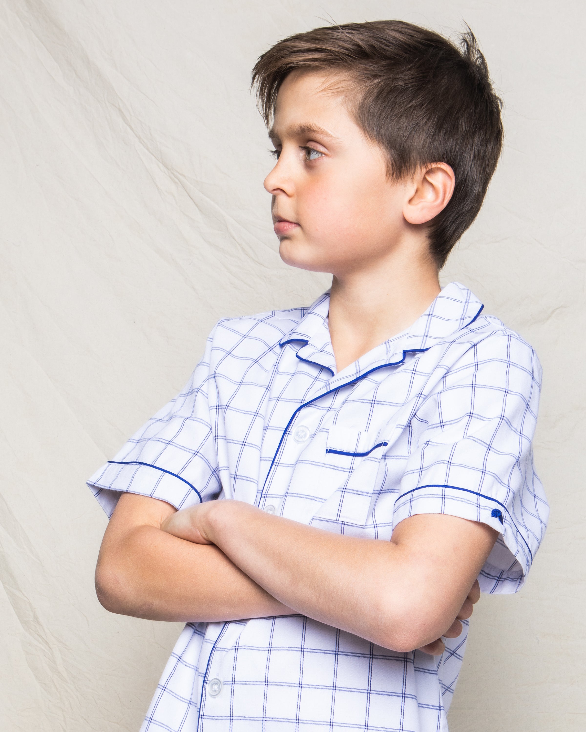 A young boy with short brown hair crosses his arms and looks to the side, wearing Petite Plumes Kids Twill Pajama Short Set in Nantucket Tattersall. The white shirt has a blue grid pattern, set against an off-white fabric background.