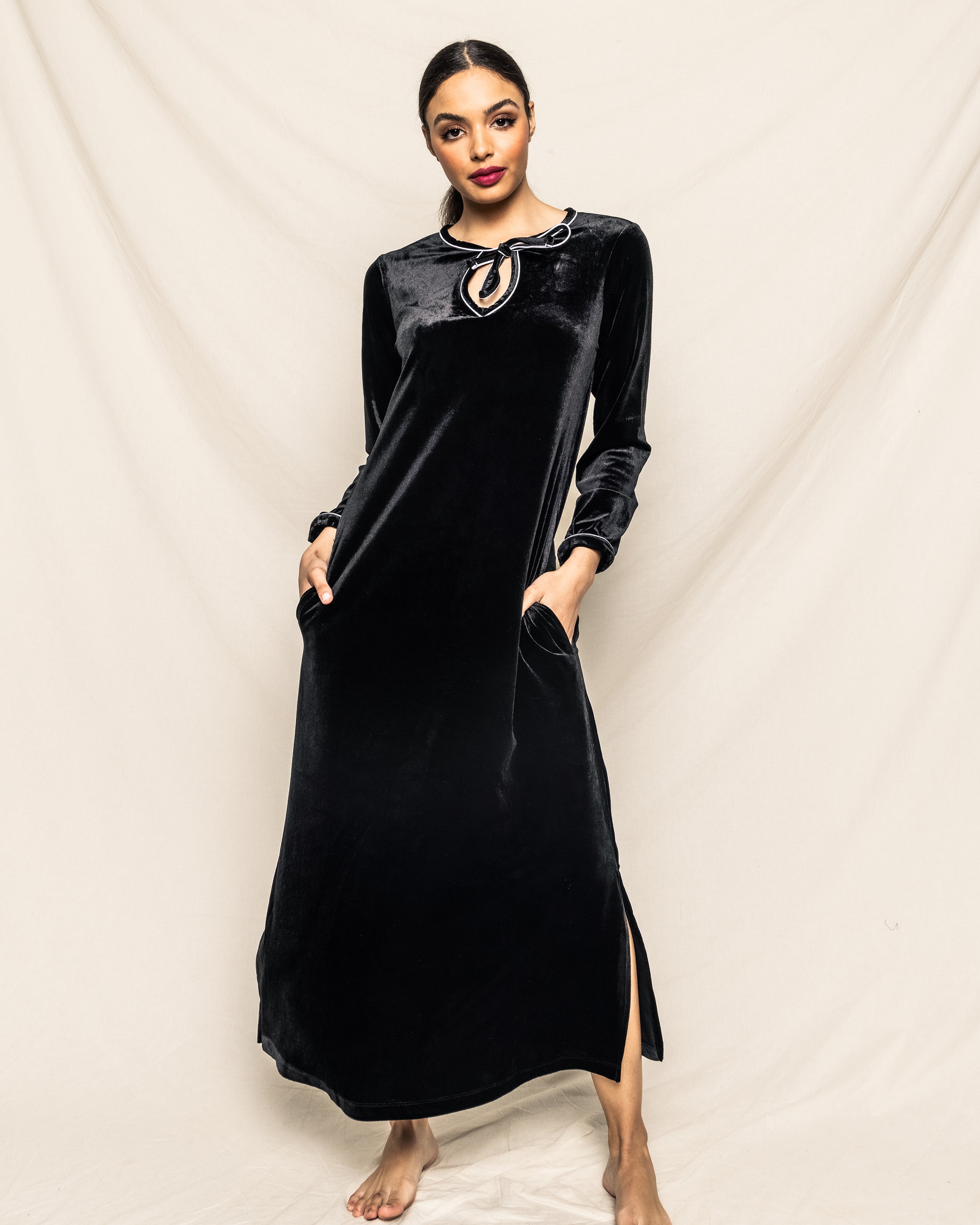 A woman stands barefoot against a light fabric backdrop, exuding classic Hollywood style in Petite Plumes Womens Velour Harlow Nightgown in black. This Cabaret Collection piece features long sleeves, a keyhole neckline, pockets, and a subtle side slit as she places her hands in the pockets.