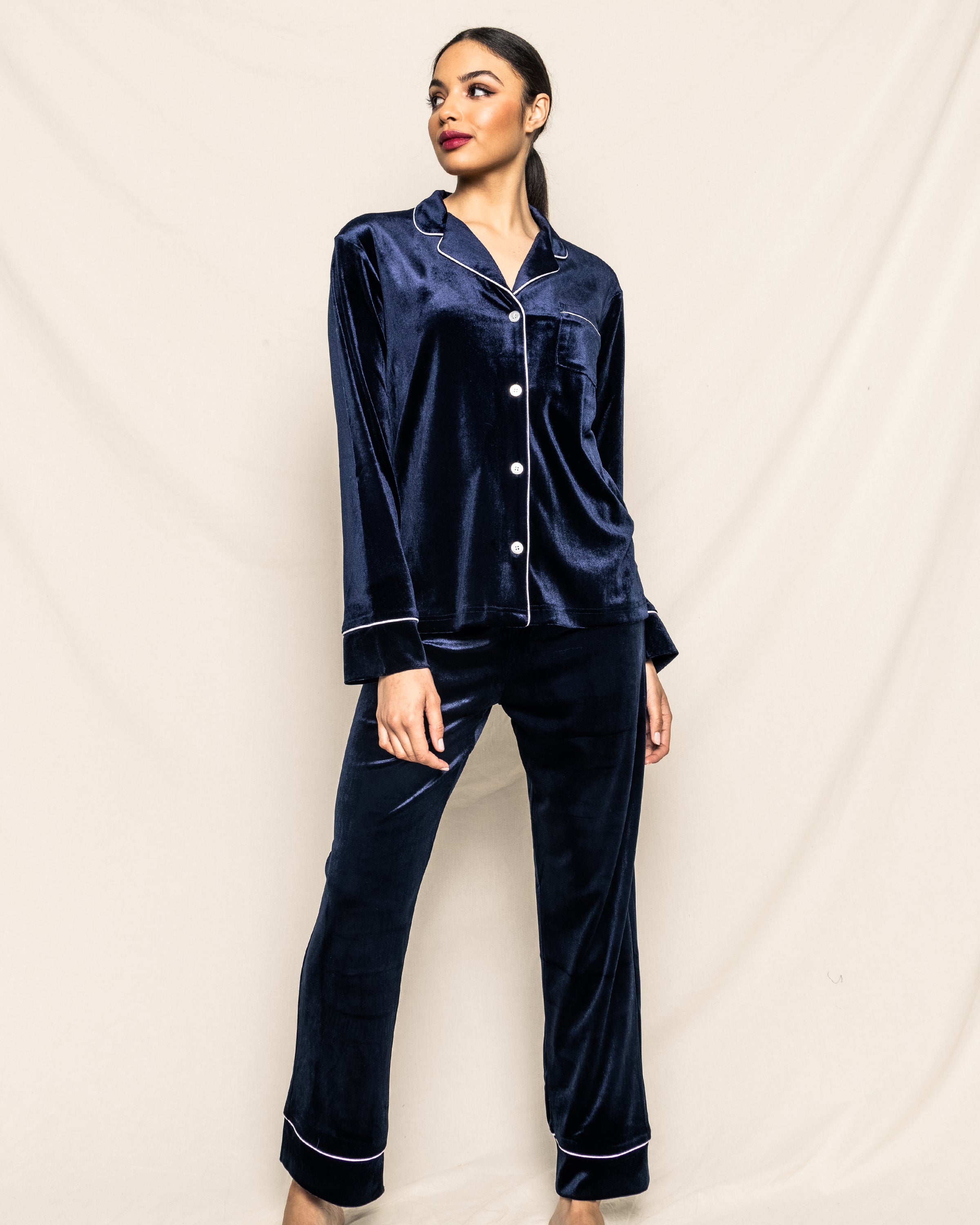 A person in a Womens Velour Pajama Set in Navy by Petite Plume from the Cabaret Collection stands against a beige backdrop, looking to the side.