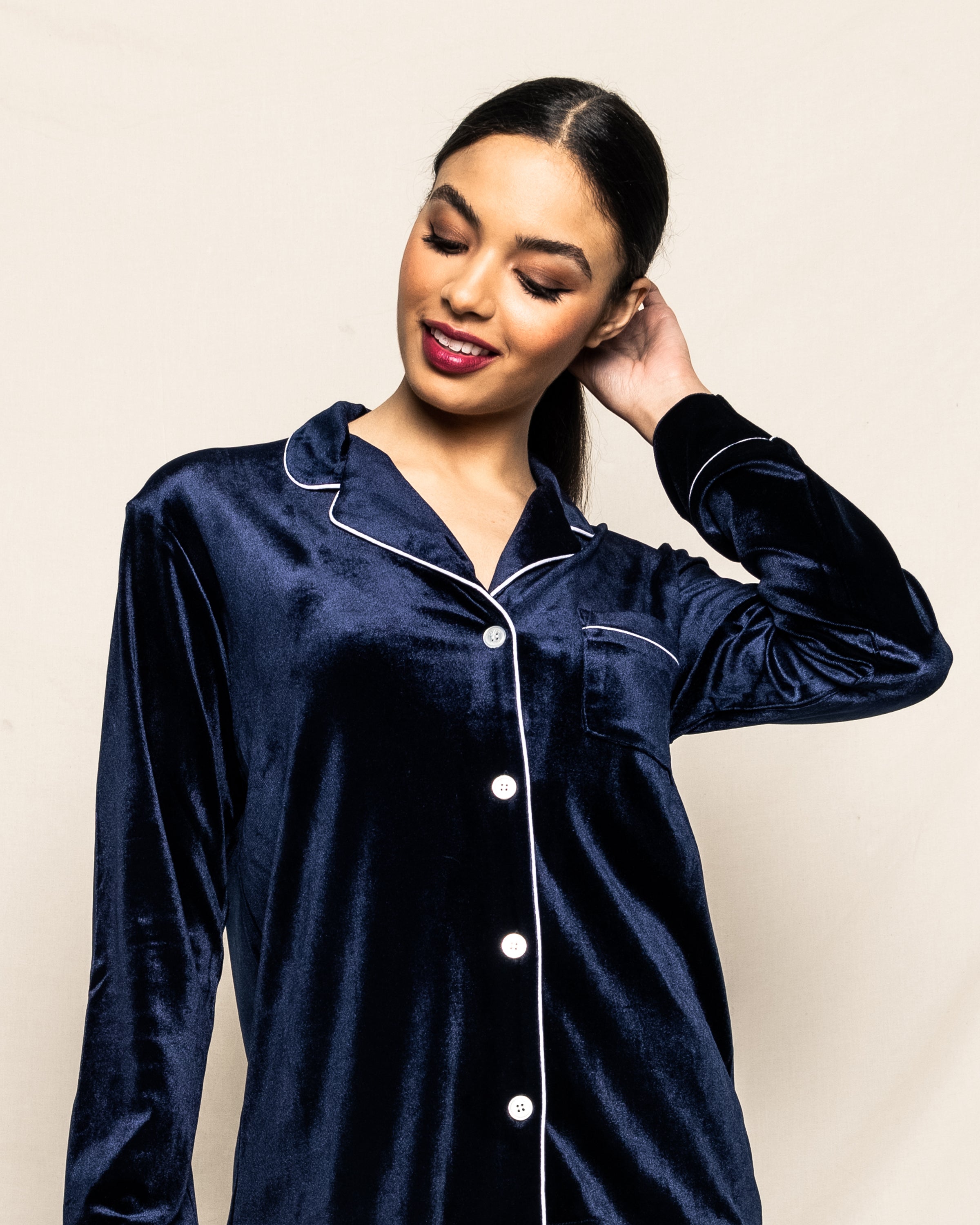 Against a plain background, a person wearing Petite Plumes Womens Velour Pajama Set in Navy with white piping, reminiscent of classic Hollywood style, smiles while touching the back of their head, their hair styled neatly back.