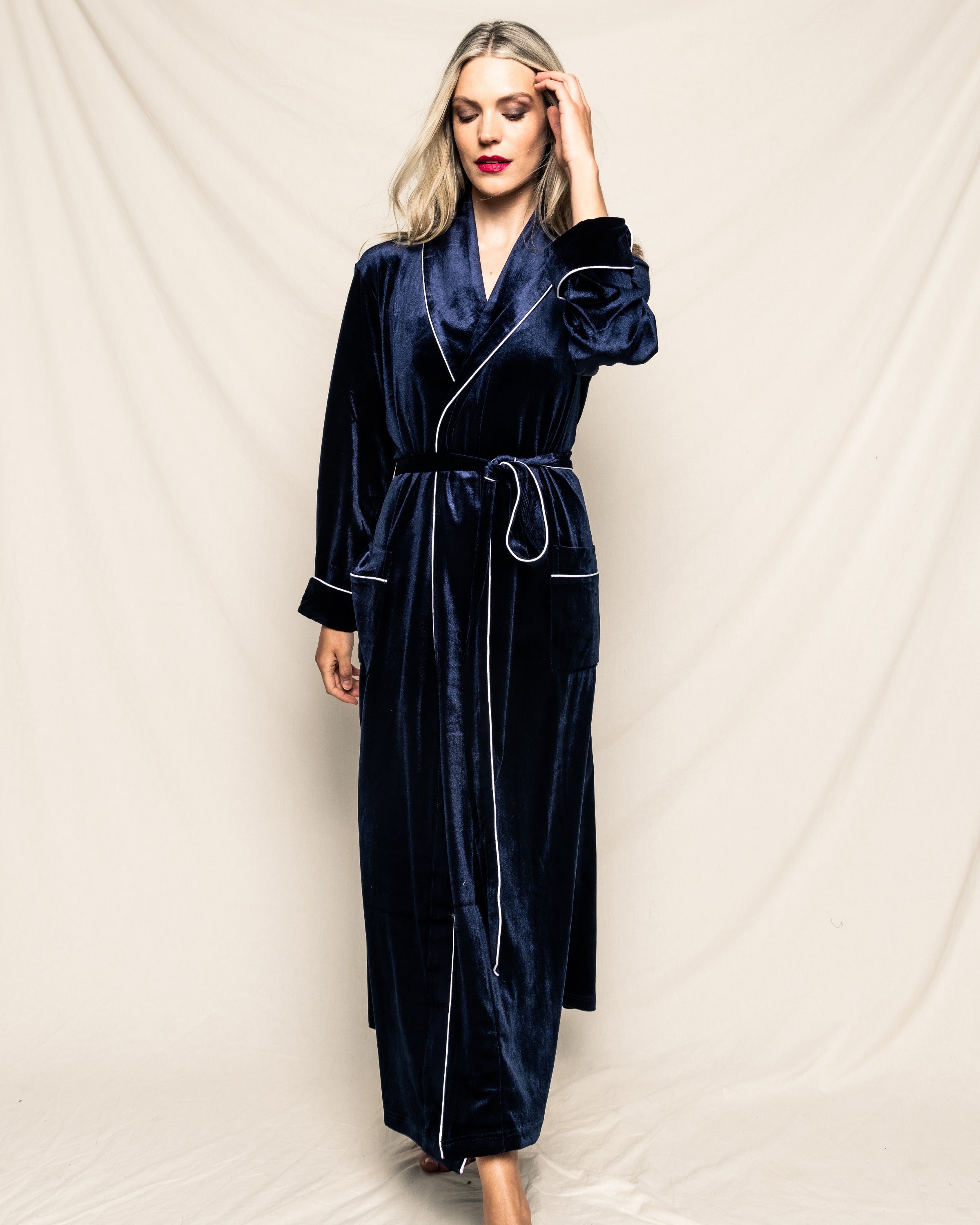 A person with long hair stands against a light beige background, wearing an exquisite Womens Velour Robe in Navy from Petite Plume. The robe features light-colored piping and a tied belt, as they look down and touch their hair with one hand.