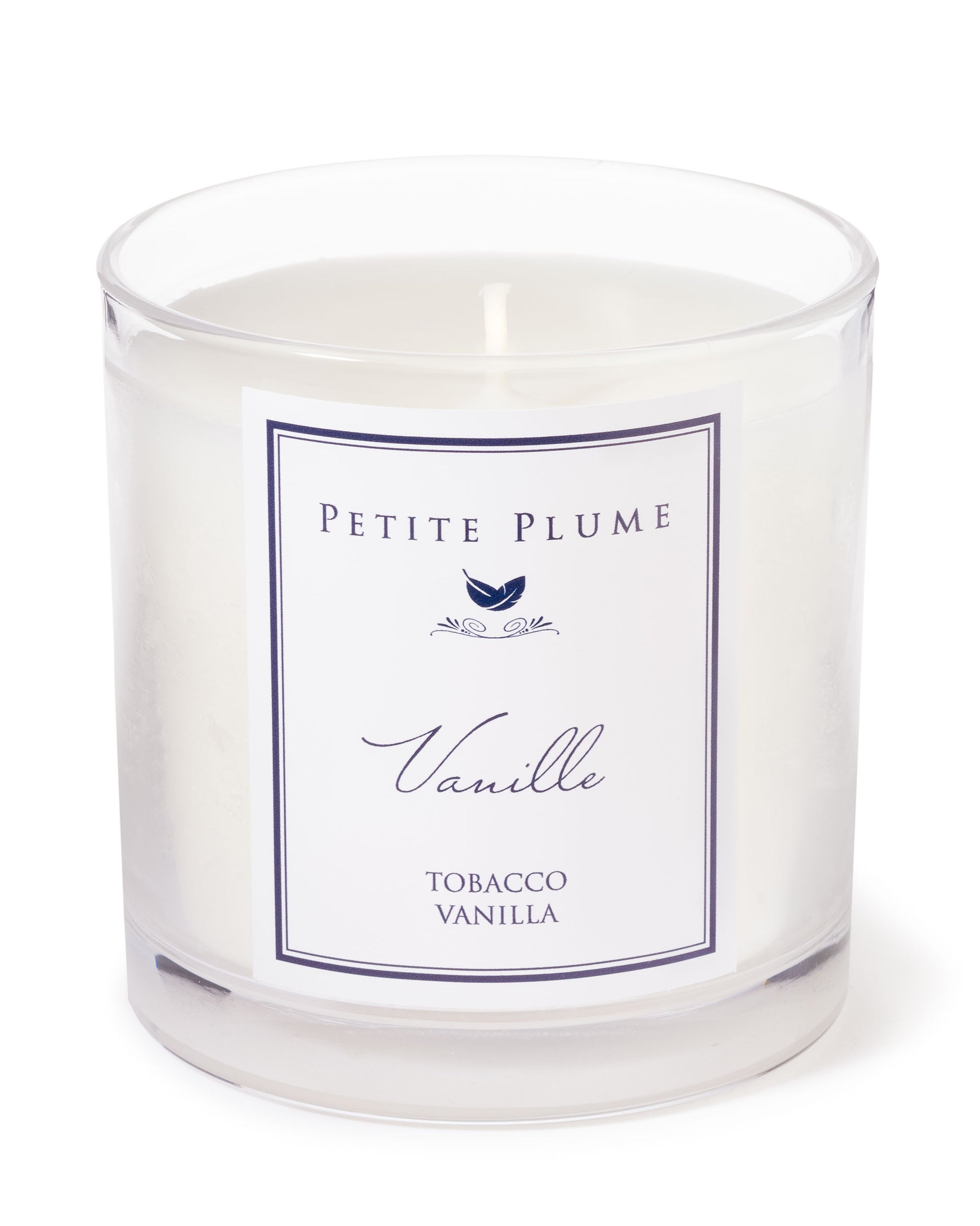A white candle in a clear jar, labeled Petite Plume Luxe Vanille Candle, exudes Vanilla Tobacco scents. Beneath the name, a charming blue bird illustration adds whimsy to this aromatic treasure from Petite Plume.